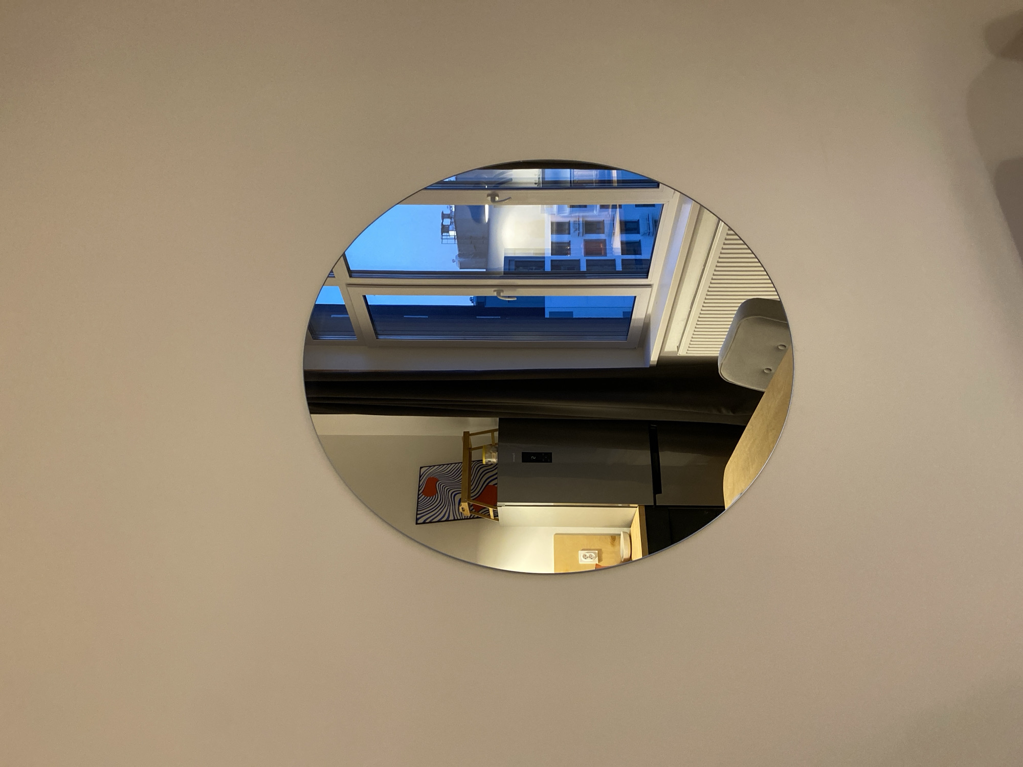 The image shows an oval mirror hanging on a light beige or light yellow wall. The reflection in the mirror reveals a room with a skylight showing a building exterior, possibly at dusk or dawn given the blue sky. The room also includes a chair, a gray refrigerator, a blue and white patterned rug, and an electrical outlet on a wooden surface. The setting appears to be indoors. The mirror's asymmetrical shape gives a distorted perspective of the reflected scene. There is no specific indication in the image to confirm that the location is Izhevsk, Russia.