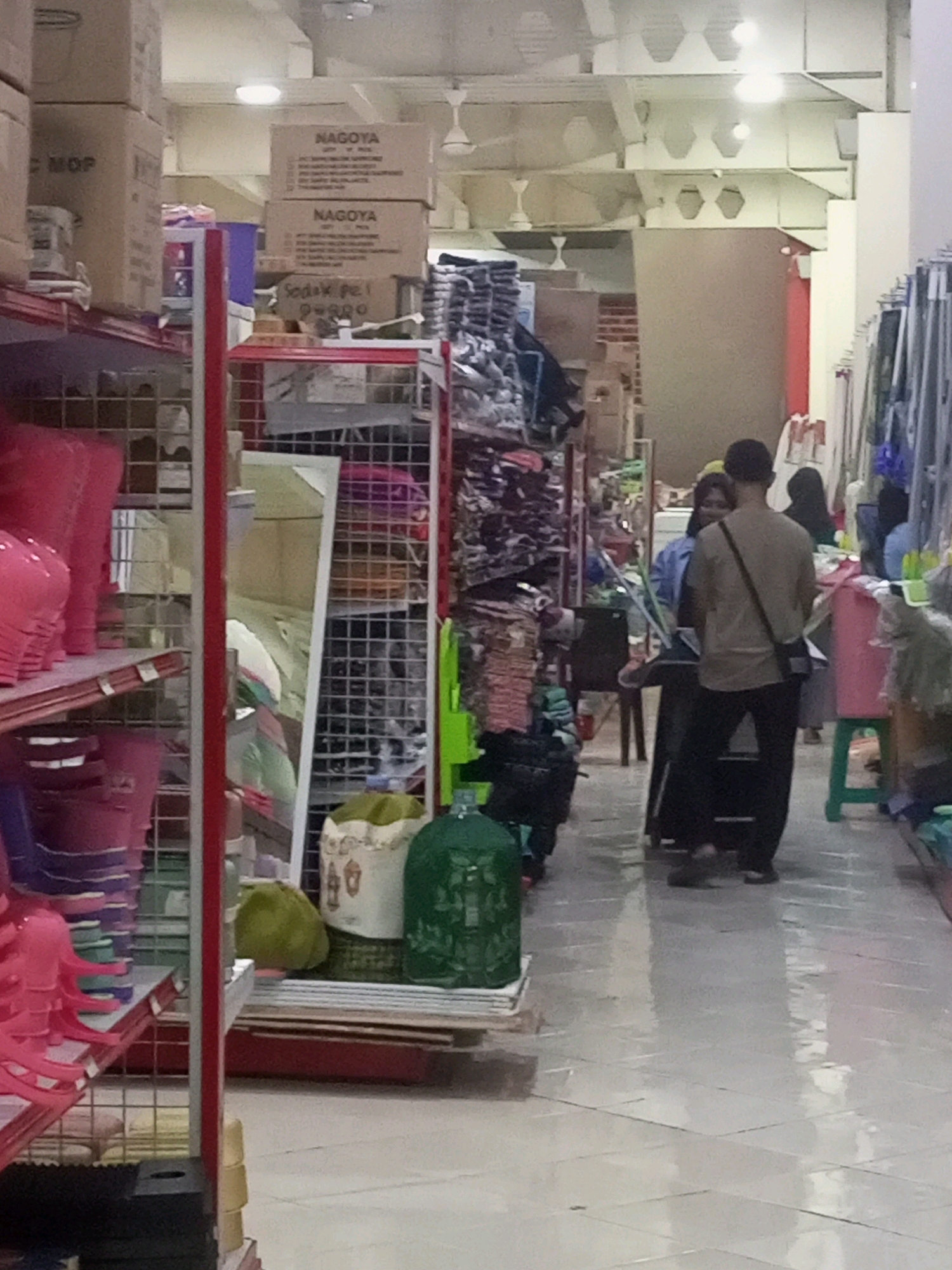 This is a medium-angle, eye-level, indoor shot of a retail store aisle in Ngasem, Indonesia. The aisle is lined with shelves on both sides, displaying various household items. On the left, pink buckets and plastic items are stacked on shelves. On the right, a man in a tan shirt and black pants, with a black bag slung across his shoulder, stands facing away from the camera. Beside him, a woman in a blue top is partially visible. Further down the aisle, there are more shelves stocked with products and several green plastic containers. The flooring is tiled with a light-colored pattern, and overhead lighting illuminates the scene. The overall impression is one of a busy retail environment.