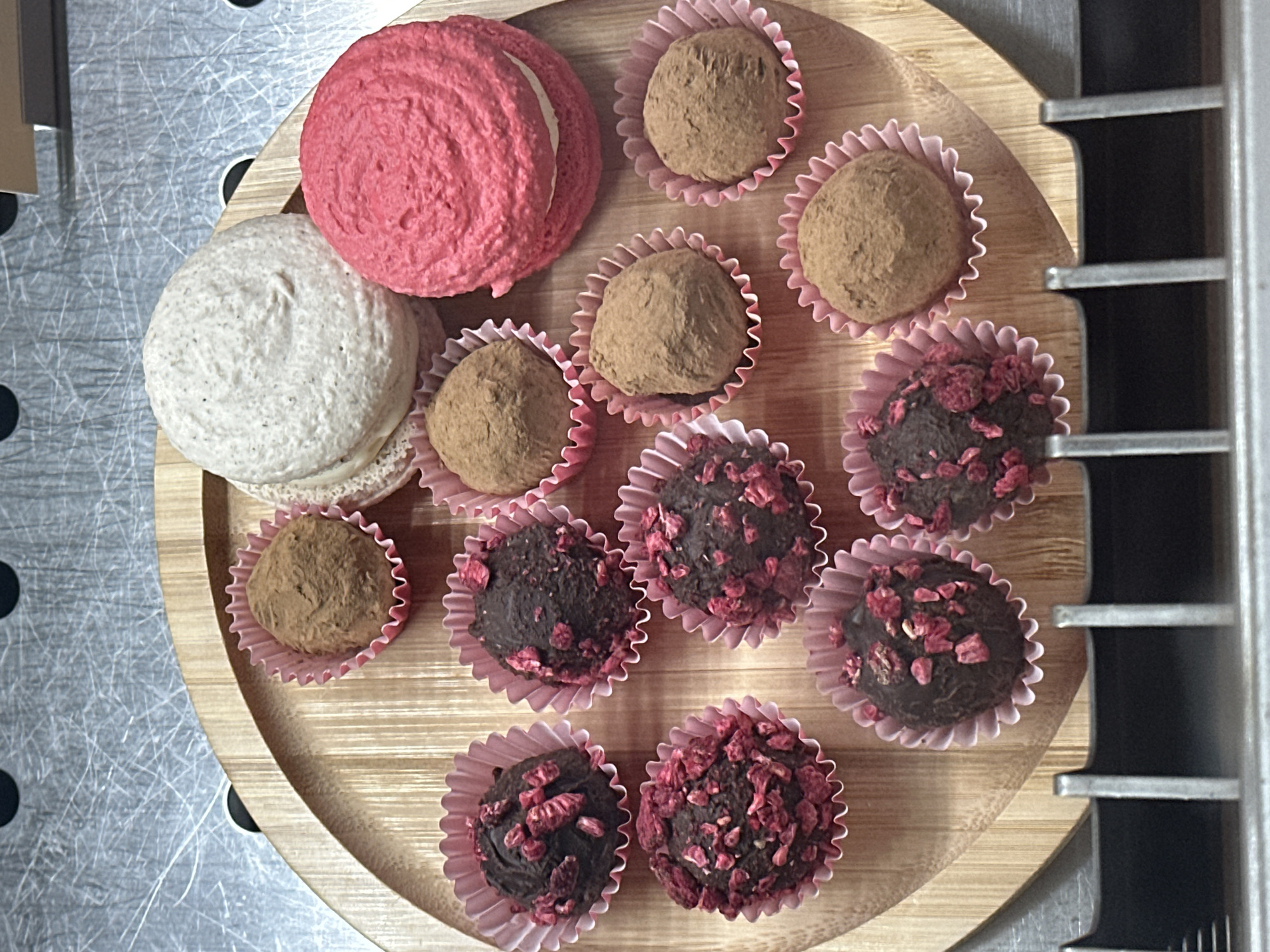 A top-down shot depicts a wooden circular tray holding an assortment of confectionery items: two macarons (one pink, one off-white) and eleven truffles in pink ruffled paper cups. Six of the truffles are coated in brown powder, while the other five are coated with what appears to be dried raspberry pieces. The tray is set on a surface with a brushed metal finish. Parts of a metal structure with horizontal bars are visible at the right. The setting is in an indoor location, potentially in Novi Sad, Serbia. There are no people depicted.