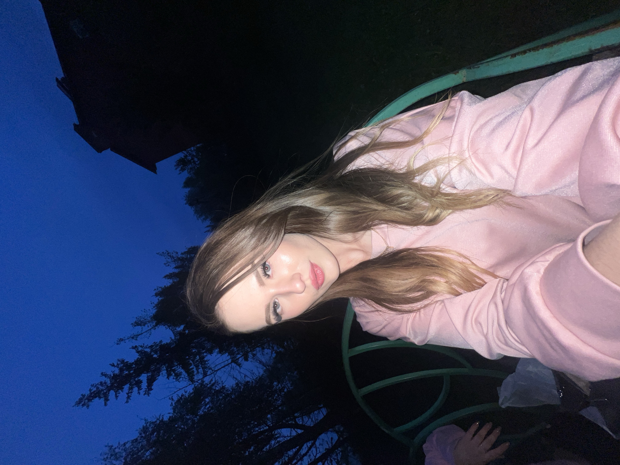 A young woman with long brown and blonde hair is lying down on a green metal structure, possibly a bench or swing, outdoors at night. She is wearing a light pink top. The background shows a dark blue sky and silhouettes of trees and a building. The location is the city of Noviy, Russia.