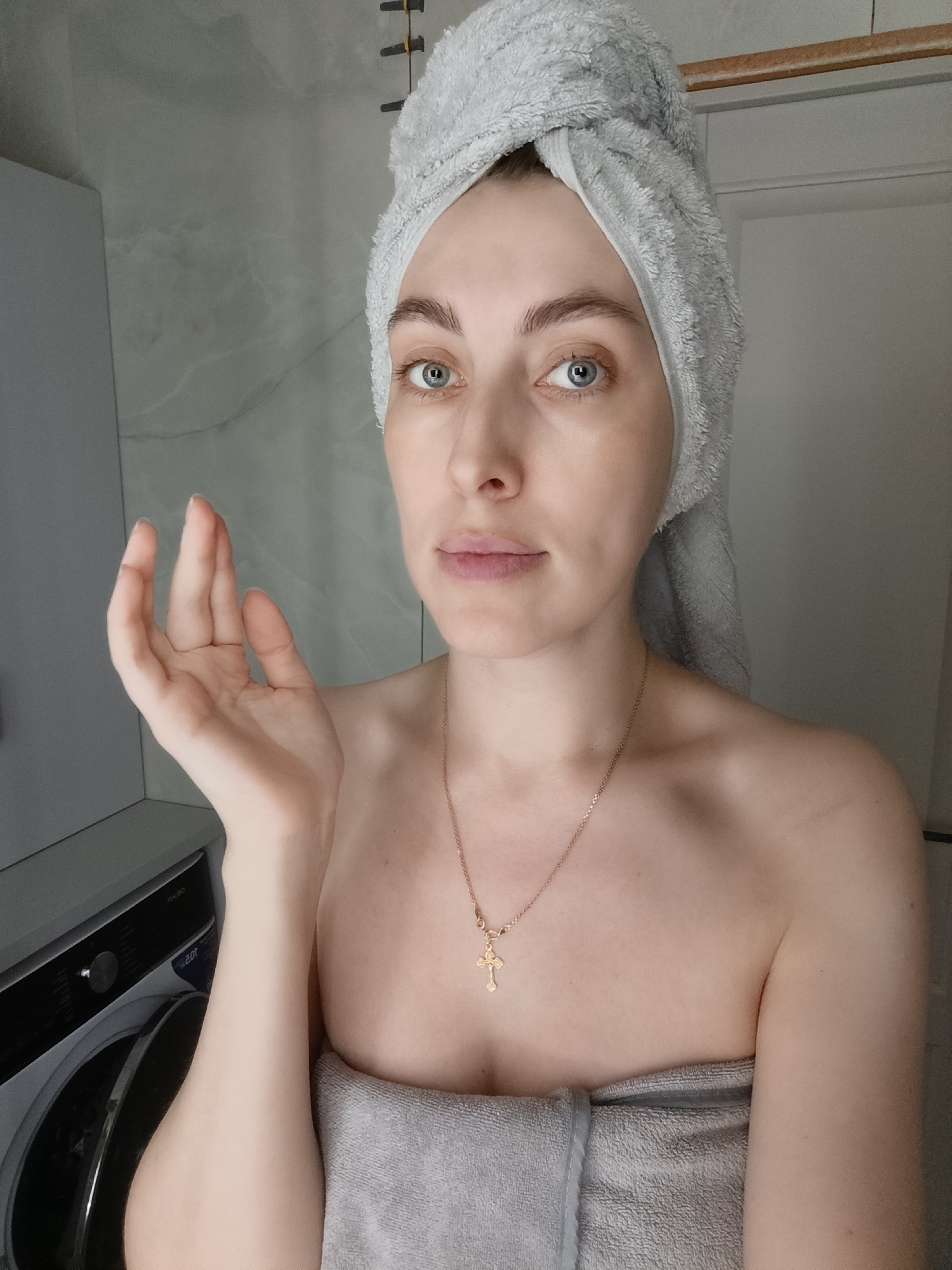 A young woman is centered in the frame, wearing a gray towel wrapped around her head and body, suggesting she is in a bathroom. She has fair skin, blue eyes, and is wearing a delicate gold necklace with a cross pendant. Her right hand is raised with fingers slightly spread. Behind her, a washing machine is visible on the left, and the wall and a door are on the right. The lighting appears to be artificial. Based on the available information and the reference to "Chaikovsky, Russia", it is possible that this photo was taken in a residential bathroom in that city.