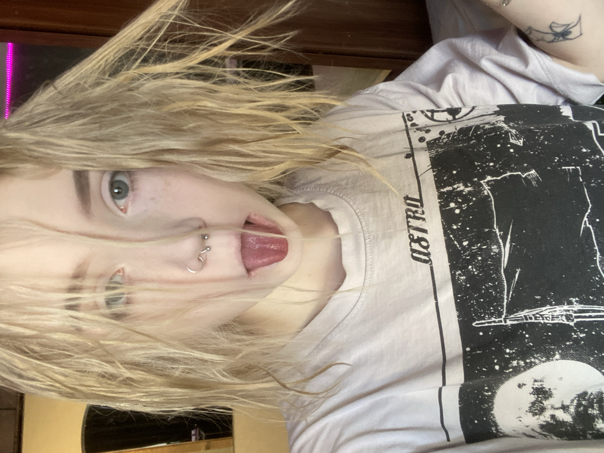 A front-facing, close-up image captures a young individual with disheveled, wet-appearing blonde hair partially obscuring their face. The subject has blue-green eyes, a light complexion with visible freckles across the nose and cheeks, and is extending their tongue. Facial piercings include a silver circular barbell in the septum, a silver hoop in the left nostril, and two small silver studs in the right nostril. The individual wears a light-colored t-shirt with a prominent black graphic print that features the word "ASTRO," a large moon, and a stylized rocket or space structure. A small, dark tattoo, appearing to be an arachnid or insect, is visible on the subject's left forearm. The background is an out-of-focus indoor environment, revealing dark wooden furniture or paneling on the right, a reflective surface with a light-colored frame on the left, and a strip of magenta LED lighting across the top right. The scene is situated in the city of Izhevsk, Russia.