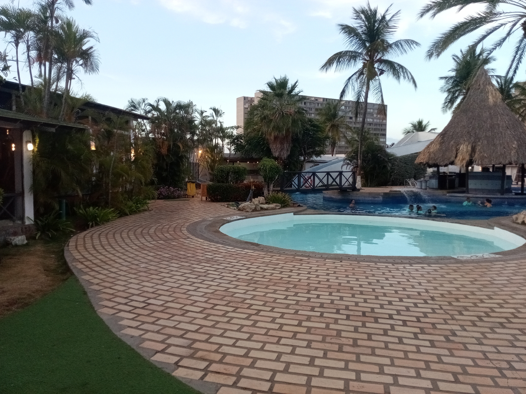 This image depicts a vibrant resort or hotel pool area in Porlamar, Venezuela, captured during the late afternoon or early evening. The scene features two swimming pools: a smaller, circular, shallow pool in the foreground, and a larger, irregularly shaped pool situated behind it.
A wide, curved paved walkway, constructed from light-colored brick-like pavers, encircles the foreground pool. A strip of green artificial turf borders this pathway on the left. The entire setting is rich with tropical vegetation, including numerous palm trees of varying heights that frame the pools and buildings.
Several individuals, including adults and children, are visible in the larger swimming pool, casually enjoying a swim or standing in the water. To the right of this pool, a distinctive thatched-roof structure, likely a bar or cabana, enhances the tropical aesthetic. A small wooden bridge with decorative cross-pattern railings spans a section of the pool area further in the background.
Buildings are prominent in the scene. On the left, a low-rise structure with a dark roof and illuminated interiors suggests a restaurant or lounge. In the mid-ground and background, a tall, multi-story building, likely the main hotel, stands against the soft, diffused light of the sky. Its facade appears somewhat aged. Artificial lights are beginning to illuminate the buildings and pathways, indicating the transition from daylight to dusk. The overall atmosphere conveys relaxation and leisure in a tropical vacation environment. No discernible text is visible within the image.