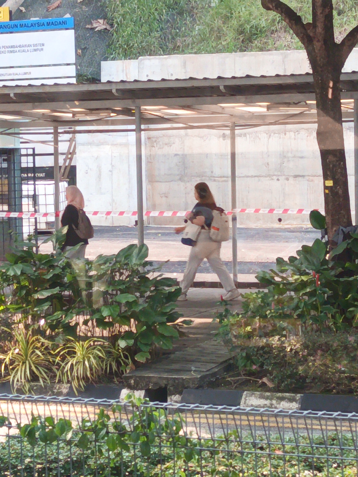 The image depicts an outdoor scene with two individuals walking along a paved pathway. In the foreground, lush green foliage and a wire fence are visible. In the background, a building with a covered walkway and a large tree are present. A sign above the walkway displays text that appears to be in Malay, with "KUALA LUMPUR" clearly legible, indicating the location is in Kuala Lumpur, Malaysia. The individuals are walking away from the viewer, one wearing a hijab and carrying a backpack, and the other with long hair and carrying multiple bags. Red and white caution tape is strung along the pathway, suggesting a construction or restricted area.
