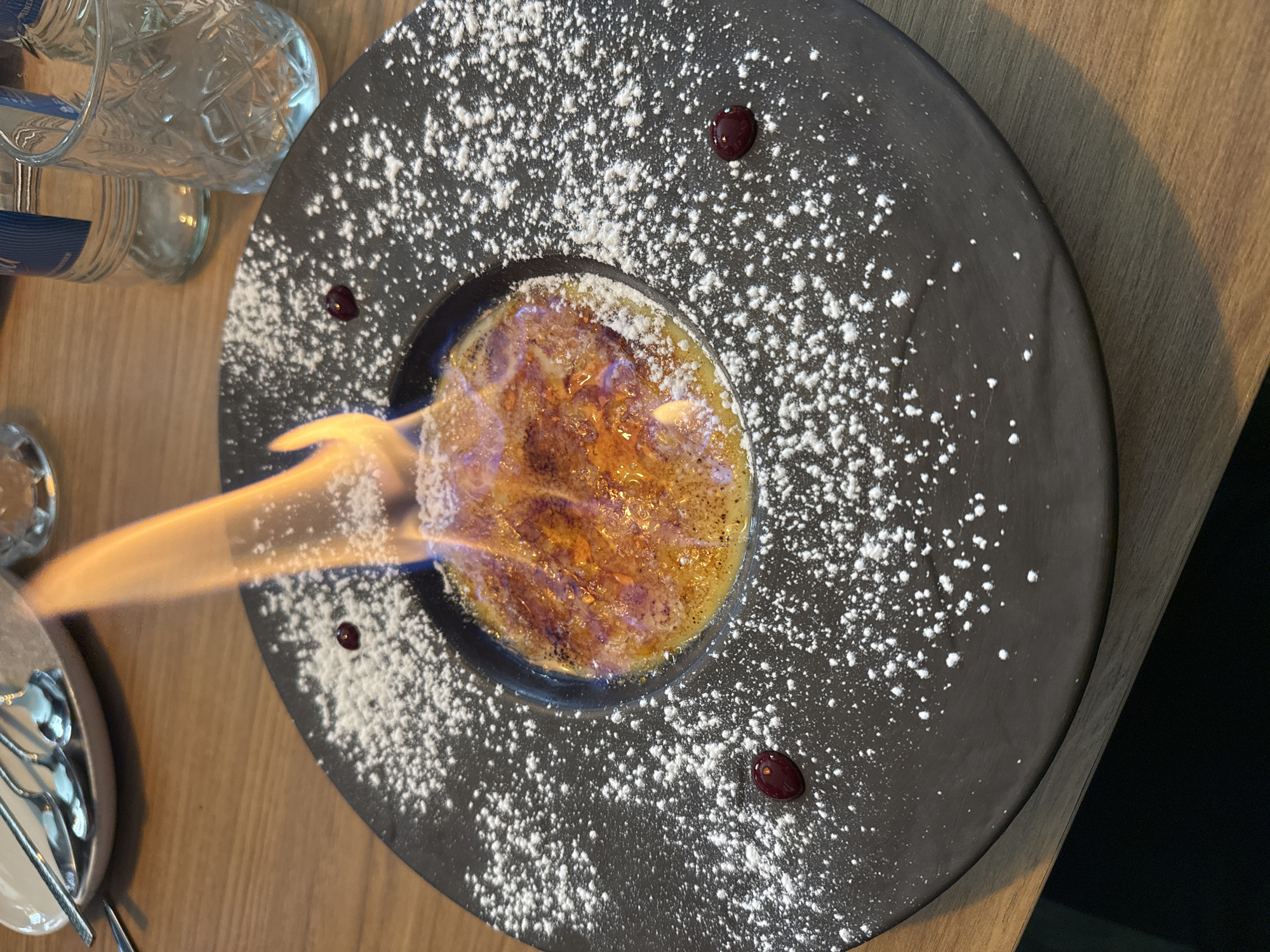 A close-up, slightly high-angle shot shows a black plate of crème brûlée being torched. The dessert sits in a shallow, black-rimmed bowl, the top caramelized to a golden-brown hue. The dark plate is dusted with a generous amount of powdered sugar, punctuated by three small, dark red fruit pieces. The flame from the torch is directed towards the dessert, emanating from the left side of the frame. The plate rests on a wooden table. The background includes a glass water bottle and cutlery in a light-colored bowl. The location is Junglinster, Luxembourg. No people are visible in the image.