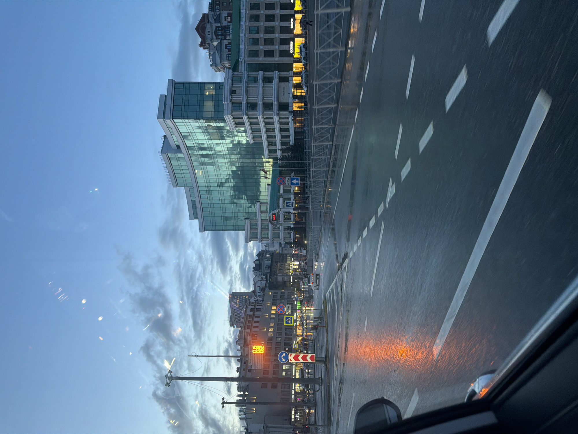 This media file captures an urban street scene in Kazan, Russia, at dusk or early evening, likely after a recent rain. The image, taken from inside a vehicle, shows a wet asphalt road with clear white lane markings and reflections of artificial lights.
In the foreground, the dashboard and side mirror of the vehicle are visible. The road surface is slick and reflects the orange glow of distant traffic lights or building lights, as well as the white of the lane markers.
The midground features a busy street lined with buildings of varying architectural styles. To the left, a modern building with a curved, reflective glass facade dominates, mirroring the cloudy sky. Further along the street, a mix of multi-story buildings, some with traditional brickwork and others with more contemporary designs, are illuminated by numerous windows and ground-floor business lights. Visible street furniture includes traffic light poles, international road signs (e.g., blue circles with white arrows indicating turns, a pedestrian crossing sign, a red circle with a diagonal line implying no stopping/parking), and a prominent red and white chevron sign for a bend or obstruction.
Several signs with Cyrillic text are visible on the buildings. One large, illuminated sign reads "АК БАРС" (AK BARS), likely a bank, with smaller text below. Another sign shows "СИРИУС" (SIRIUS). A digital display on one building appears to show the time, possibly "18:00".
The sky above is overcast with patches of soft blue and grey clouds, suggesting the transition from day to night. The overall atmosphere is quiet and reflective, characteristic of an urban evening following precipitation. No people are distinctly visible in the frame.