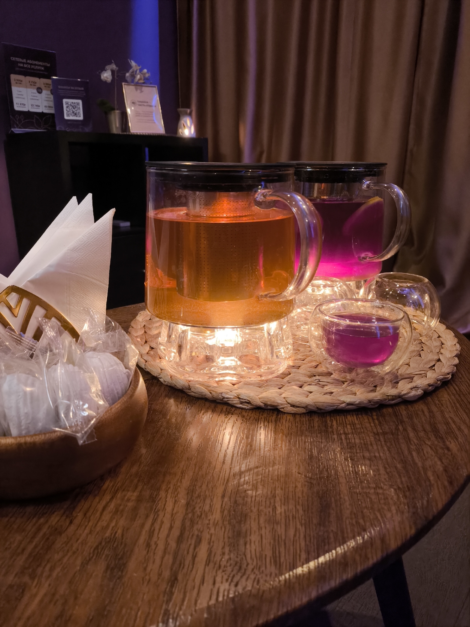 On a round wooden table in a dimly lit space is a setting with two glass mugs filled with colorful beverages, one orange and the other pinkish-purple, sitting on a woven straw placemat along with several small glass cups. To the left, a wooden bowl holds wrapped candies and a stack of white napkins. In the background, there is a black shelf with decorative items and signs, and draped curtains. Given the Russian text on the sign, the location is likely Moscow, Russia. There are no people in the scene. The beverages are the primary subjects. The table, cups, and candies are the objects. The scene is a still life setup.