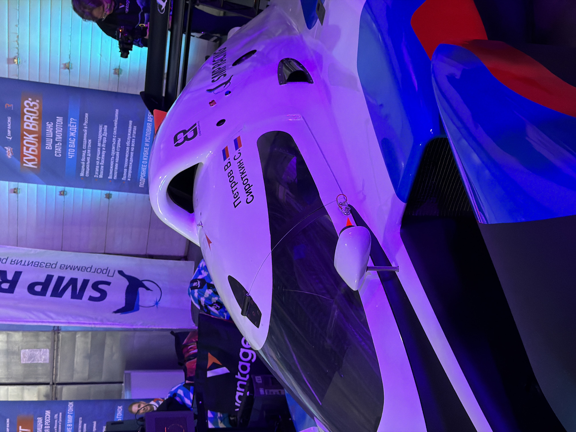 Here is a detailed description of the image:
**General Description:**
The image is a close-up shot of a race car's front section, possibly taken at an event or exhibition. The car features a predominantly white body with blue and red accents and black details. The lighting appears to be predominantly purple, casting a distinct hue over the scene.
**Objects & Subjects:**
- **Race Car:** A white, blue, and red race car with visible aerodynamic features like the nose cone, side mirrors, and possibly a front splitter. Names "Петров В." (Petrov V.) and "Сироткин С." (Sirotkin S.) are written on the car, likely identifying the drivers. The number 8 is also visible on the vehicle.
- **Signage:** Various banners with text. One prominently displays "SMP RACING" and "КУБОК BRO3:" (KUBOK BRO3:). Other texts on the banners describe the competition, including details about "Moscow Raceway" and "Игора Драйв" as race locations.
- **Background elements:** People are visible in the background, including one appearing to hold a camera. There's also a banner with "SMP R" visible, along with other indiscernible items.
**Actions & Interactions:**
- The main action is implied by the presence of the race car and event signage. It suggests an event focused on racing, competitions, or exhibitions related to SMP Racing and KUBOK BRO3.
**Environment:**
- The setting appears to be an indoor exhibition hall or event space with lighting systems.
- The text mentions Moscow Raceway and Igora Drive, two well-known racetracks in Russia, suggesting that the event is related to these locations.
**Location in Khimki, Russia:**
Based on the clues within the signage, the image likely pertains to a racing event or exhibition related to SMP Racing and KUBOK BRO3 which is connected to the Moscow Raceway, which is in Khimki.