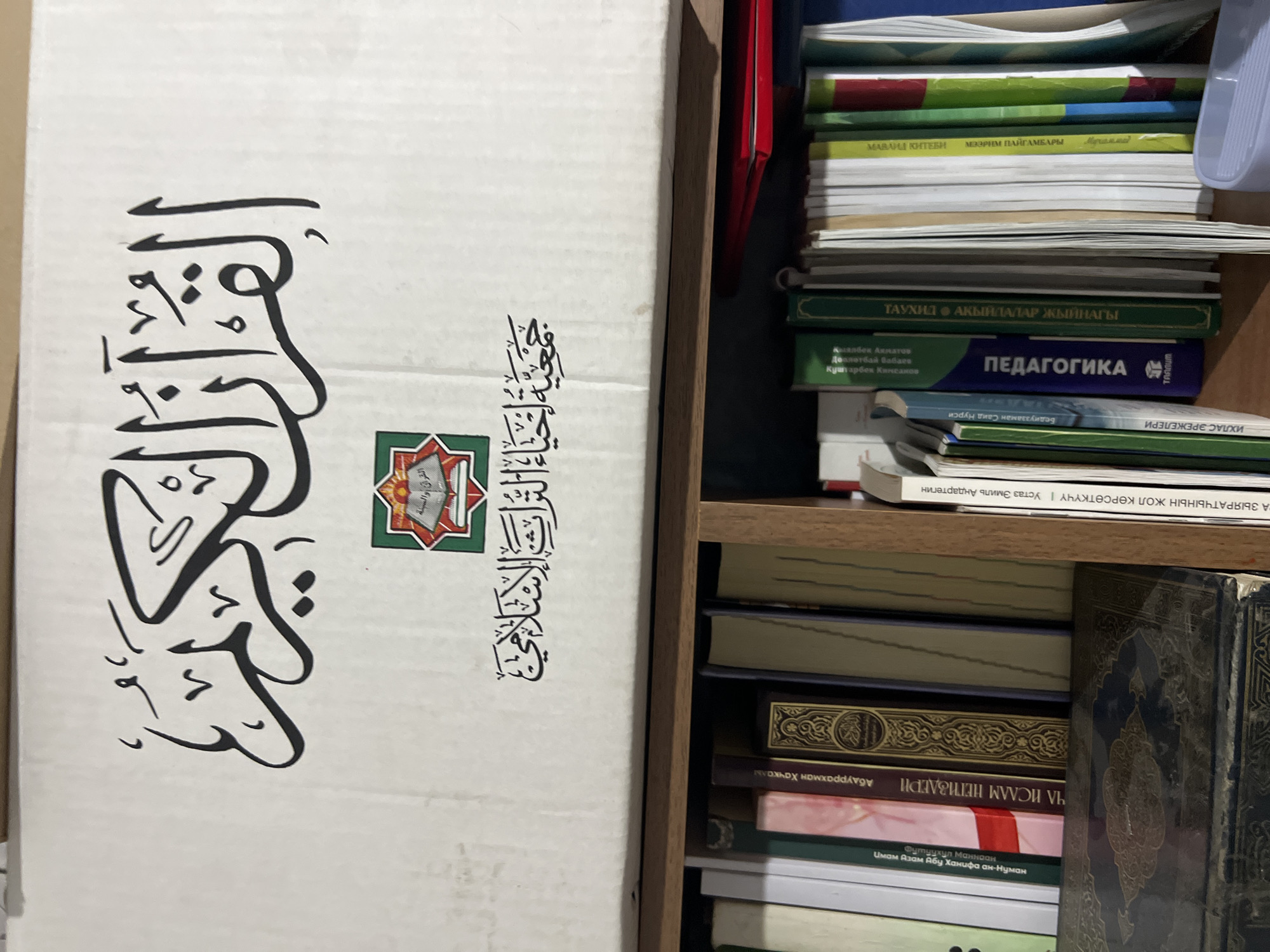 The image shows a close-up view of a cardboard box with Arabic calligraphy on it, placed above a bookshelf filled with books. The calligraphy on the box translates to "The Noble Quran" and "Society for the Revival of the Islamic Sciences." The box is white with black Arabic script. Below the box is a wooden bookshelf containing numerous books of various sizes and colors. Some book spines are visible, with titles in Cyrillic and possibly other languages. The lighting suggests an indoor setting. The scene does not provide any specific information or visual cues that would allow for the determination of a location within Bishkek City, Kyrgyzstan.