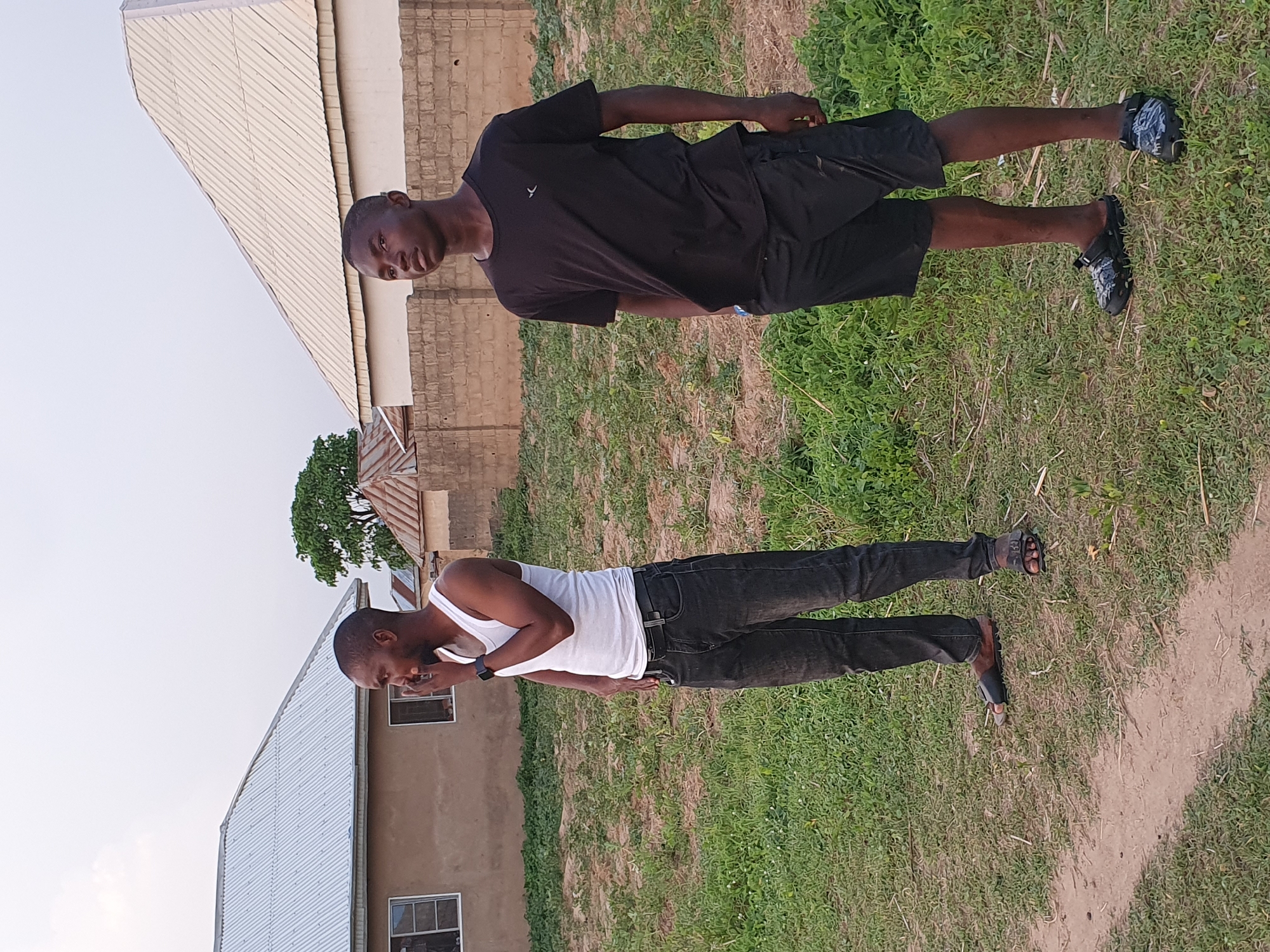 Two young Black men stand outdoors in Bana Stream, Nigeria. One wears a white tank top and black jeans and appears to be talking on the phone, while the other wears a black t-shirt and black shorts. They are both standing on a grassy area next to buildings. One building is covered in corrugated metal, and the other is made of bricks. A dirt path is visible in the grass.
