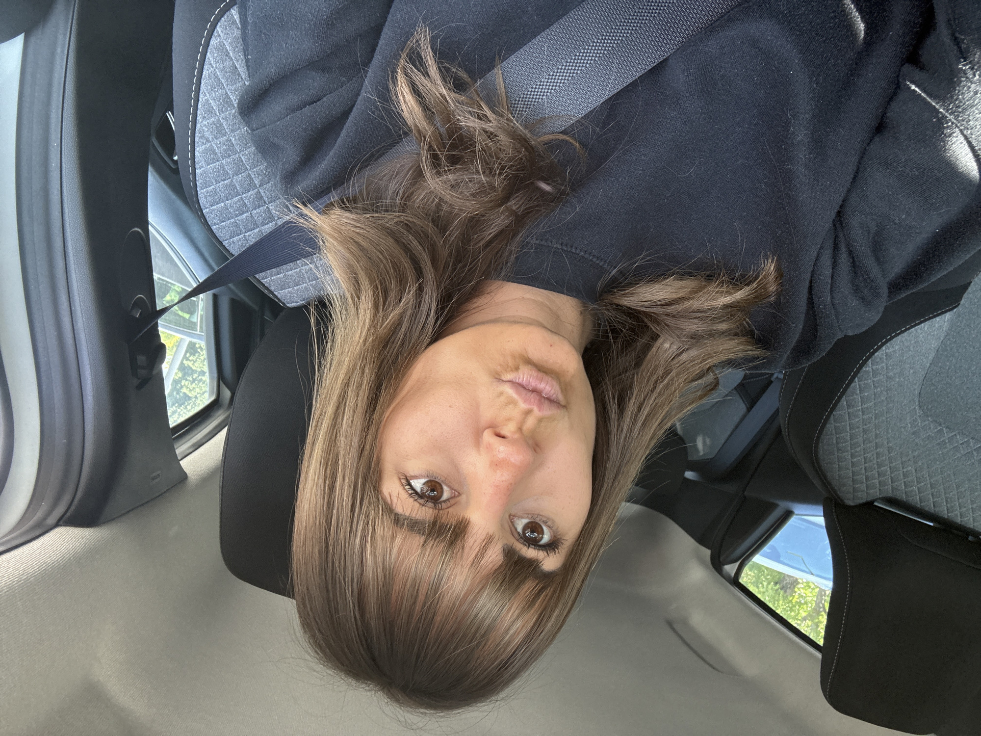 A woman with brown hair and bangs is in the driver's seat of a car. She is wearing a black hooded sweatshirt and a seatbelt. The car's interior is gray and black. A window shows a glimpse of green trees and a blue sky. The image provides no evidence to determine if she is in the city of Orange, France.