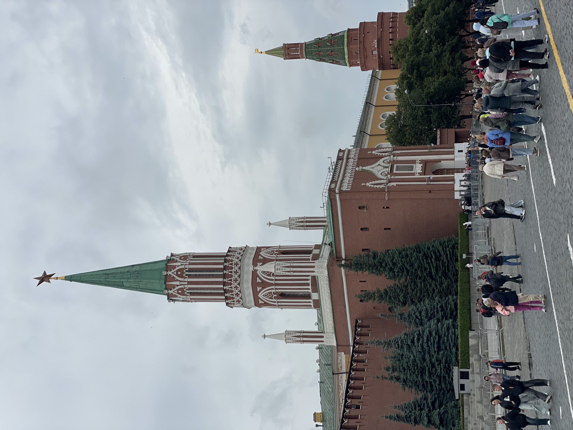 The image shows a view of the Spasskaya Tower of the Moscow Kremlin and a portion of the Kremlin Wall, with a crowd of people in the foreground. The Spasskaya Tower is tall and slender, with a green spire topped by a red star. It is made of red brick and features white decorative elements. To the right, another red brick tower with a green roof is partially visible. The Kremlin wall is also made of red brick. In front of the wall are several dark green evergreen trees. The ground is paved with cobblestones. A number of people are walking and standing in the paved area, some individually and some in small groups. The sky is overcast with gray clouds. The overall scene depicts a public space in Moscow, likely Red Square, with a focus on the iconic Kremlin architecture and a gathering of tourists or locals.