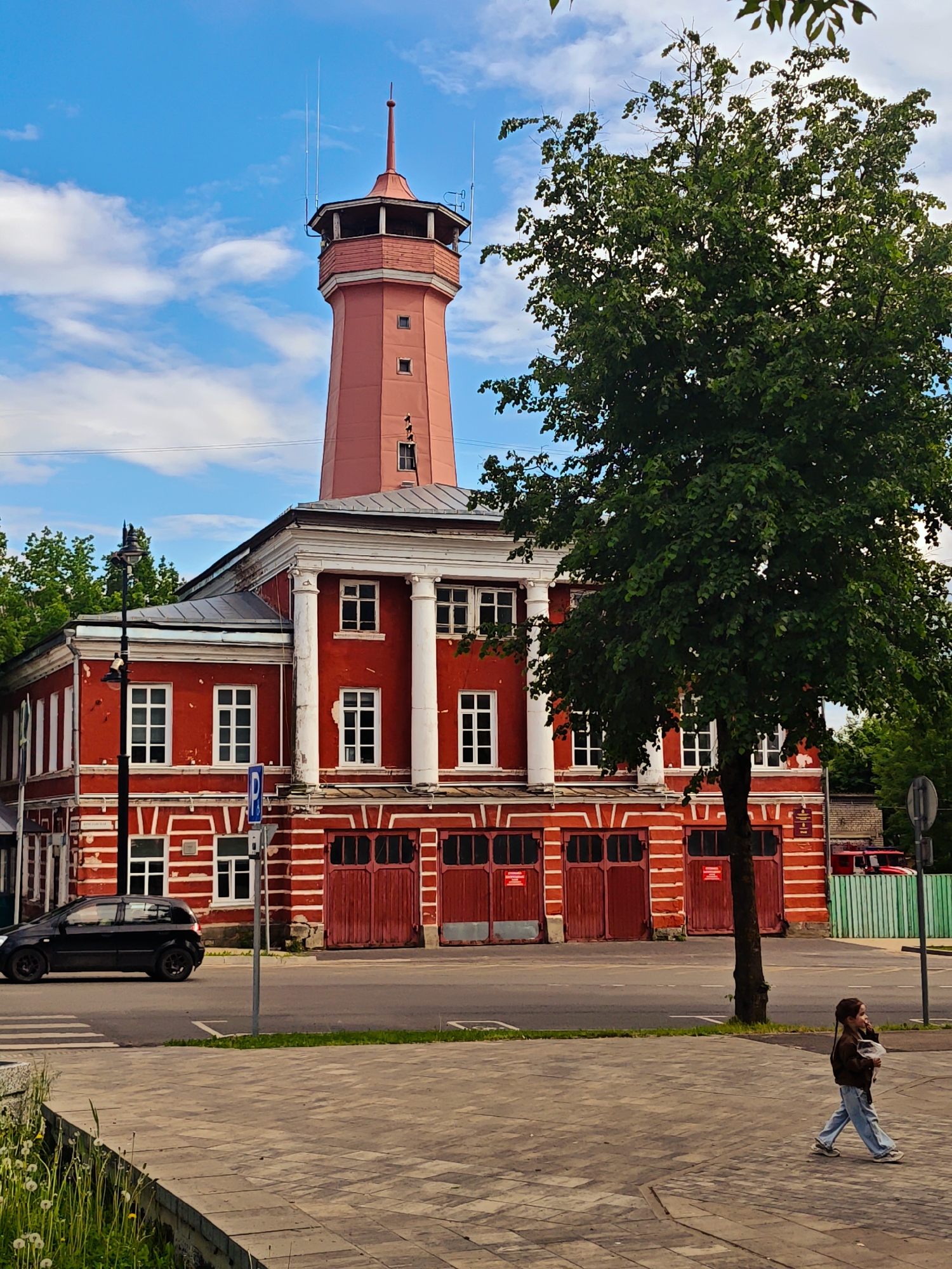 The image depicts a red and white building with a prominent red tower, identified as a fire station located in Selivanovo, Russia. The building has large white columns at the front and multiple garage-style doors. A large green tree partially obscures the building. A black car is driving on the road in front of the building. A girl walks on the sidewalk in the foreground. The sky is blue with scattered clouds. A street light stands near the sidewalk. Street signs are visible near the building.