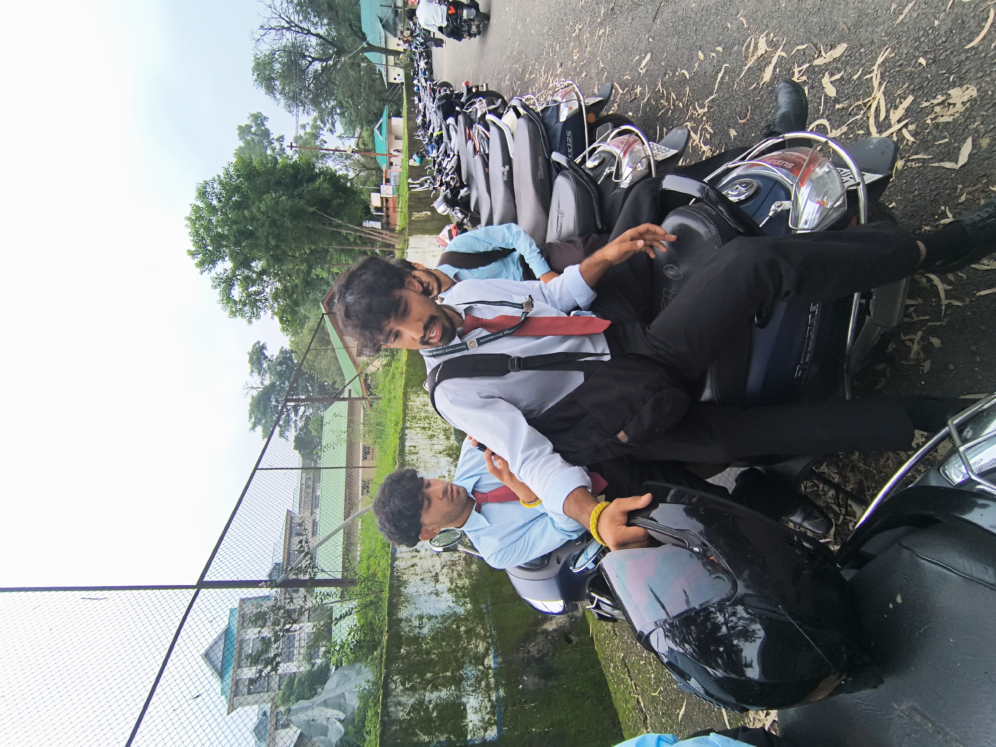 In Dharamshala, India, three young men, likely students based on their attire and the presence of scooters, are gathered around parked scooters. The foreground features a person in a light blue shirt and red tie, seated on a dark-colored scooter, holding a black helmet. Another person in a similar light blue shirt and red tie is positioned behind and to the left, looking at a mobile phone. A third individual in a light blue shirt and a backpack is partially visible to the right. The scooters are parked in a line on an asphalt surface, with scattered dry leaves. In the background, a chain-link fence encloses an area with greenery and buildings. The sky is overcast.