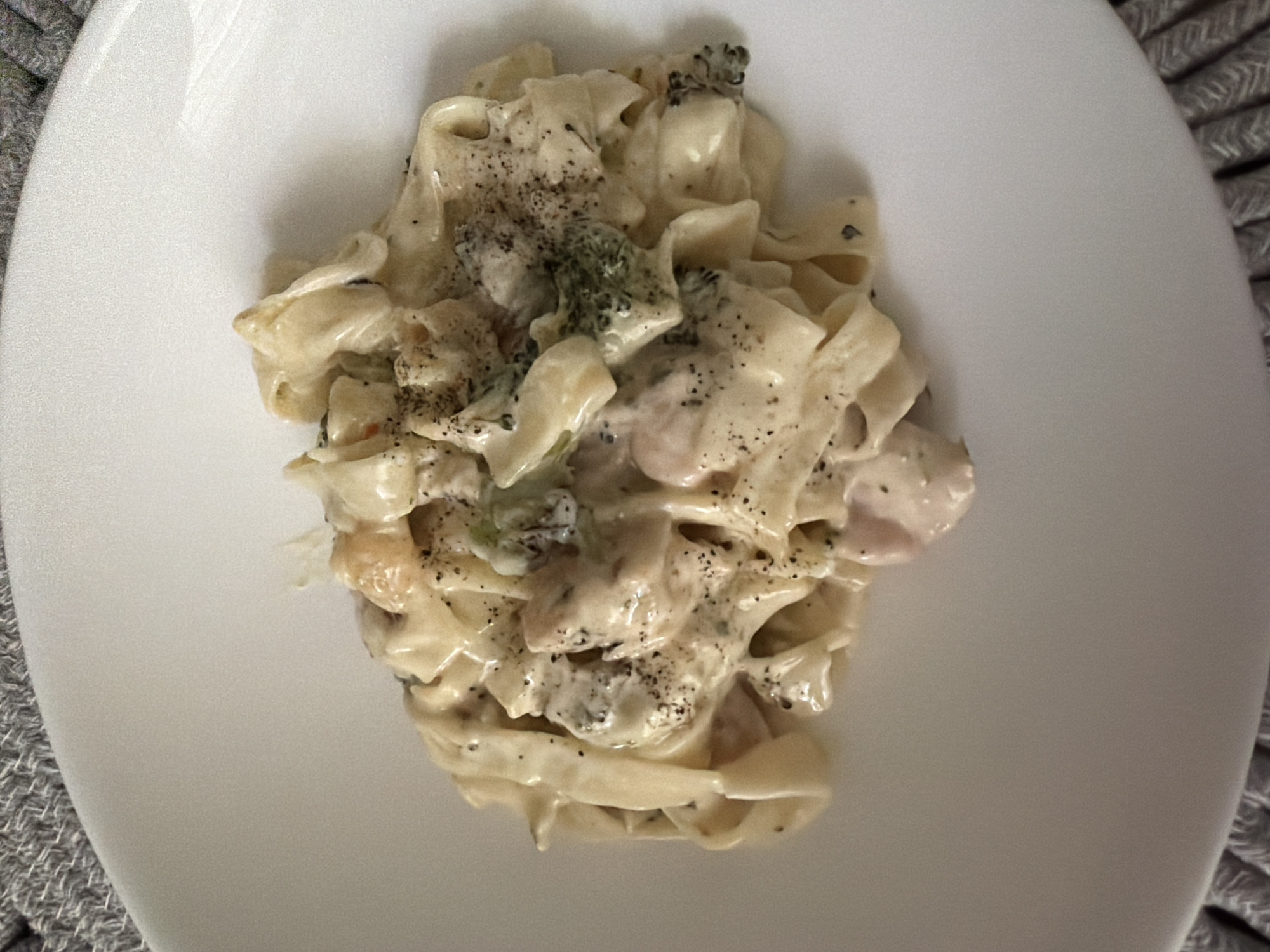 A close-up, overhead shot captures a portion of a white plate holding a serving of fettuccine pasta with chicken and broccoli in a creamy sauce. The pasta is coated in the sauce and appears to be seasoned with black pepper. Visible ingredients include pieces of chicken and florets of broccoli. The plate is set on a textured, gray placemat. The image is shot from a high angle, focusing on the food. There is no information available about the location being in Pazardzhik, Bulgaria.