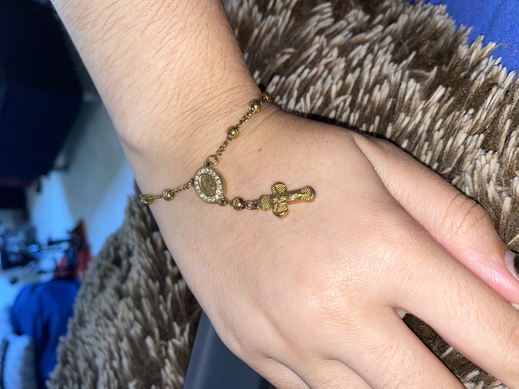 A person's hand is adorned with a gold-colored rosary bracelet. The bracelet features small, round beads connected by a delicate chain, an oval medallion with a depiction inside, and a small cross. The skin on the hand is fair. The background includes a brown, textured surface, possibly a shaggy rug or blanket, and out-of-focus elements of an interior setting, including what appears to be a blue garment and indistinct objects suggesting a room setting. There is no information to relate the image with the city of Cotoperíz, Venezuela.