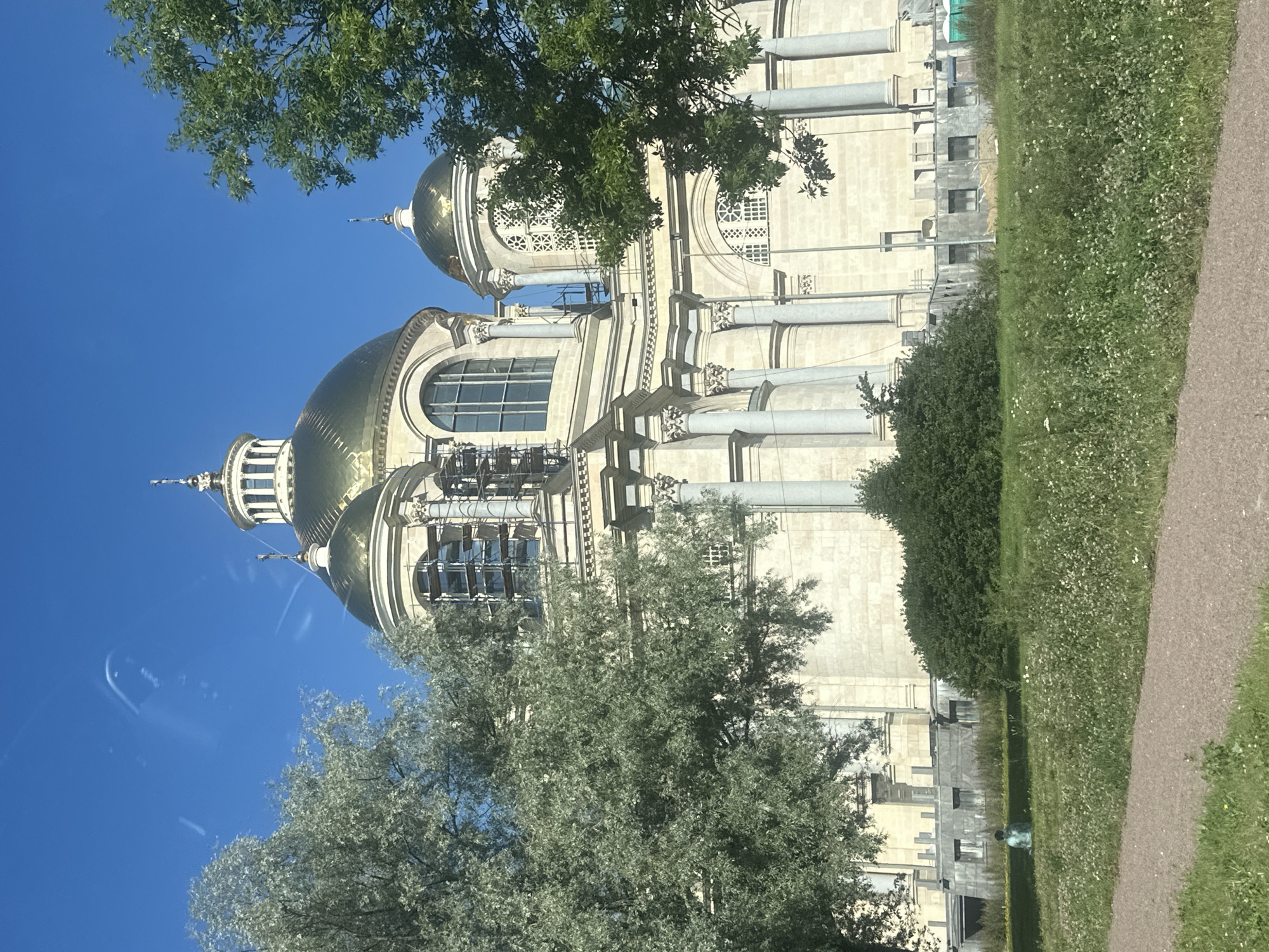 The image shows a wide-angle shot of the Saint Nicholas Naval Cathedral in Kronstadt, Saint Petersburg, Russia, on a bright, clear day. The cathedral, with its prominent golden domes and classical architectural features, is partially obscured by lush green trees in the foreground. The building is constructed of light-colored stone and features large arched windows, some of which are undergoing renovation with scaffolding visible.
In the middle ground, a grassy area with scattered white flowers stretches towards the cathedral. A solitary figure, appearing to be a person sitting cross-legged, is visible on the grass to the left of the frame. The immediate foreground shows a gravel path bordering the grassy area. The sky above is a vibrant blue with a few faint, wispy cloud formations. The overall impression is one of a sunny day in a serene, historic setting.