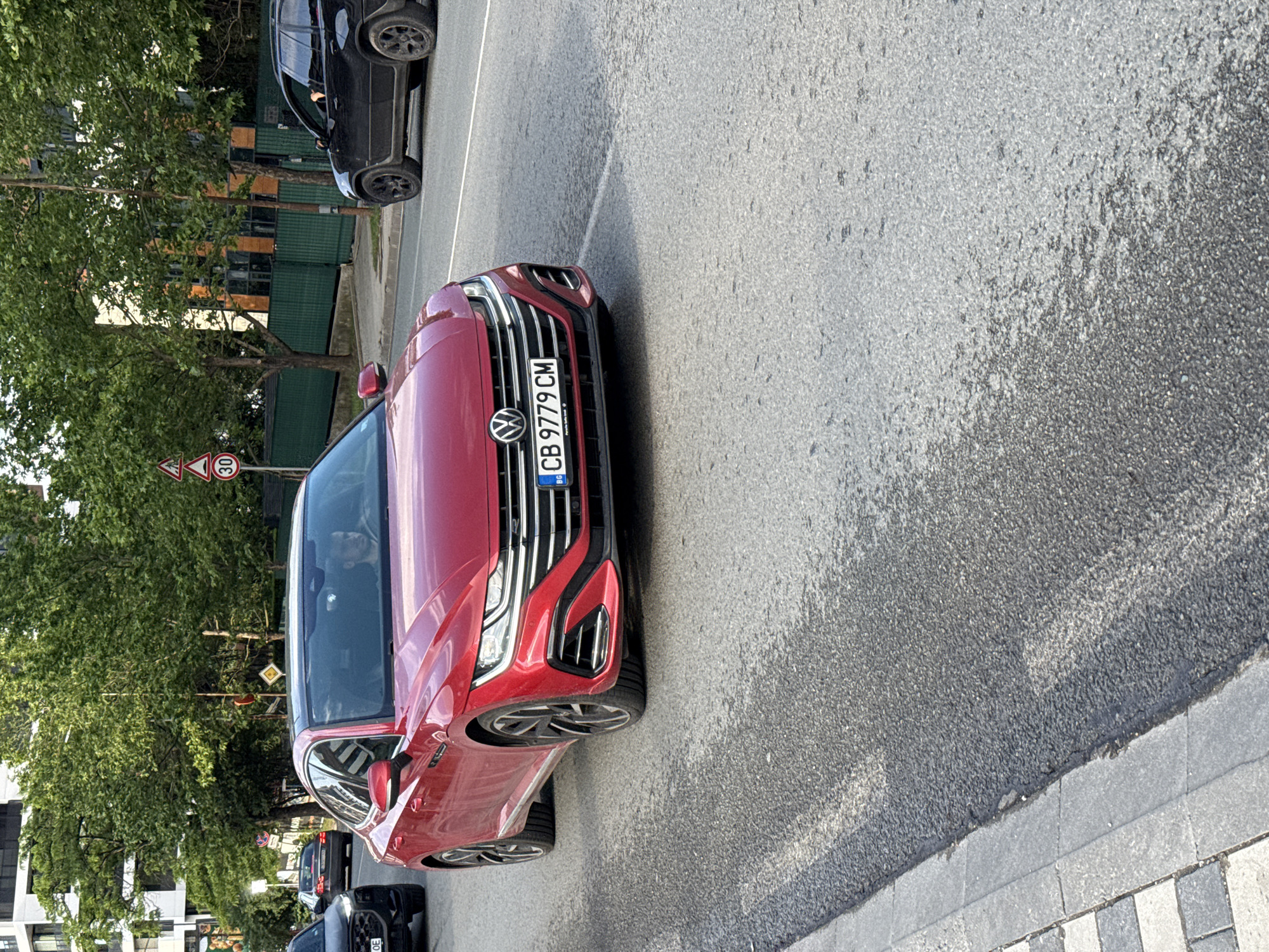 The image displays a red Volkswagen car, likely an Arteon, parked on the side of an asphalt road. The car faces slightly to the left, revealing its front grille, headlights, and a Bulgarian license plate "CB 9779 CM". The "CB" designation confirms the location as Sofia, Bulgaria, as provided in the context. The license plate holder also shows "Volkswagen" and "Porto Cars OOD".
Behind the red car, a black SUV is also parked. Another black car is partially visible in the upper left corner, suggesting a line of parked vehicles. To the left of the road, lush green trees line the street, behind which modern buildings with windows are visible.
Several road signs are present: a white circular sign with a red border and "30" in the center indicates a 30 km/h speed limit. A yellow diamond-shaped sign with a white border, commonly indicating a main road, is also partially visible.
The scene appears to be set during daytime, with clear weather and soft light, possibly early morning or late afternoon, creating some shadows on the road surface. The road itself is asphalt, and a paved sidewalk with alternating light and dark gray square tiles runs alongside it. A faint reflection of a person can be seen in the red car's windshield.