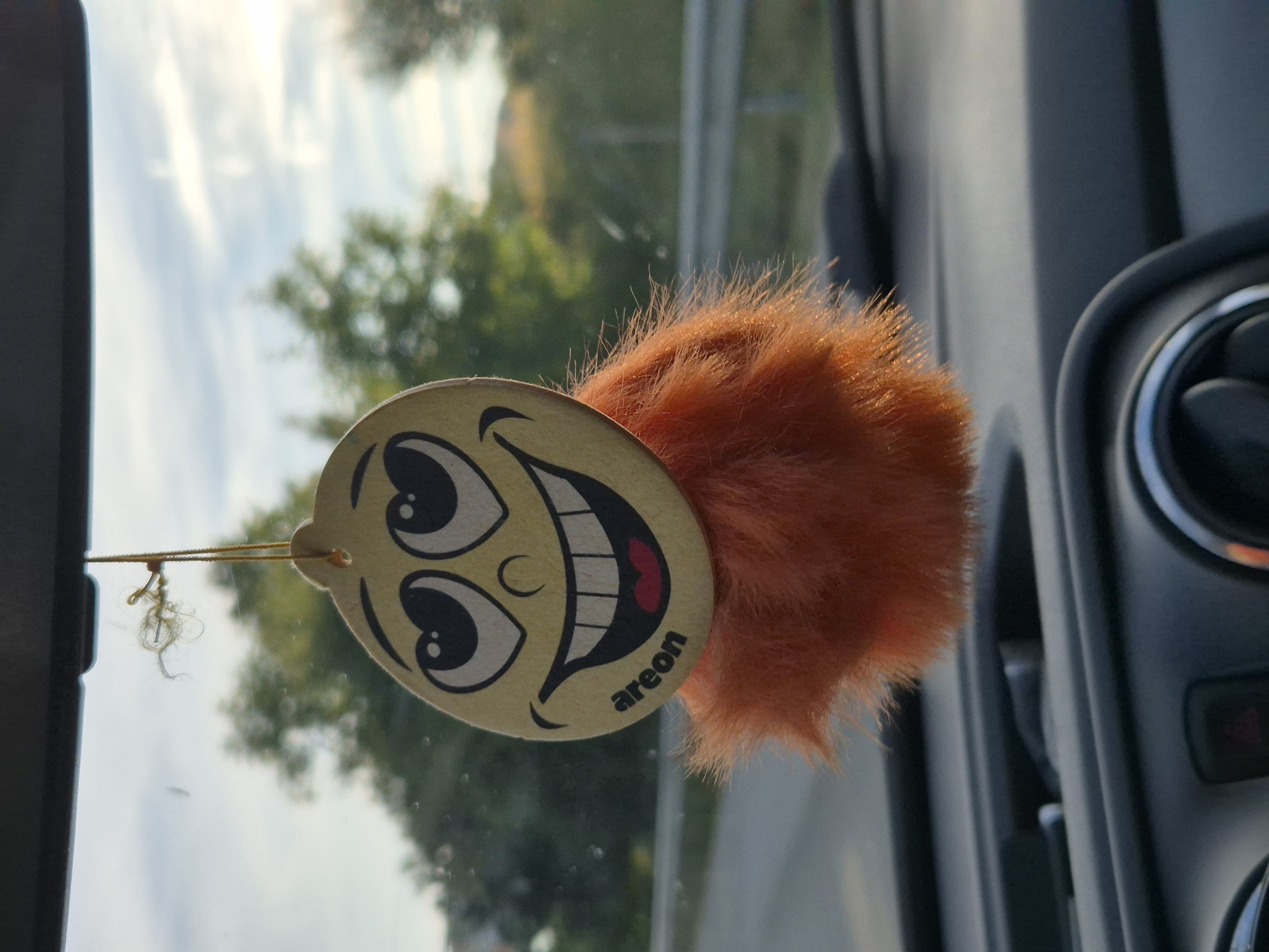 This media file shows a close-up of a car air freshener hanging from a string. The air freshener is circular with a cartoonish smiley face. The face has large, wide eyes, a wide grin showing teeth and a pink tongue, and a small circular nose. The air freshener has a fluffy, orange pom-pom attached to it. The brand name "areon" is visible in small, black text on the bottom right of the smiley face.
The air freshener is hanging in what appears to be a car's interior, with a portion of the car's dashboard and an air vent visible on the right side of the frame. The background, seen through what is likely the car's windshield, shows a bright sky with some clouds and green trees, suggesting it is daytime and the weather is fair. The overall impression is one of a cheerful and possibly scented vehicle interior. The location context of Osikovitsa, Bulgaria, does not provide specific visual cues within the image itself. The image captures a moment of everyday life inside a vehicle.