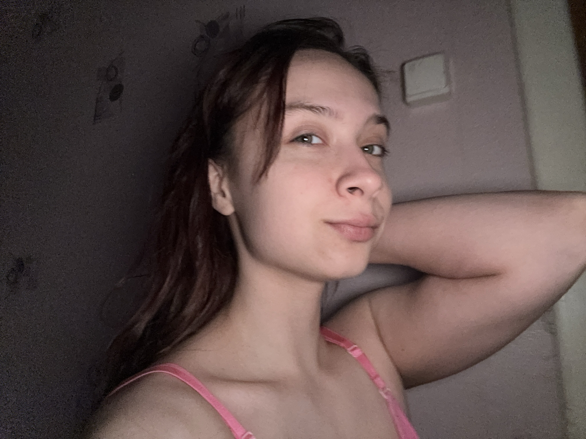 A fair-skinned young woman with brown hair, partially framing her face, looks at the camera. She has green eyes, a small nose, and thin lips. She is wearing a pink bra. Her right arm is raised behind her head. The background consists of a greyish-purple wall with dark smudges and a light switch in the upper right corner. The photograph was taken indoors, in what appears to be a bedroom. Location information is extraneous and cannot be determined from the image.
