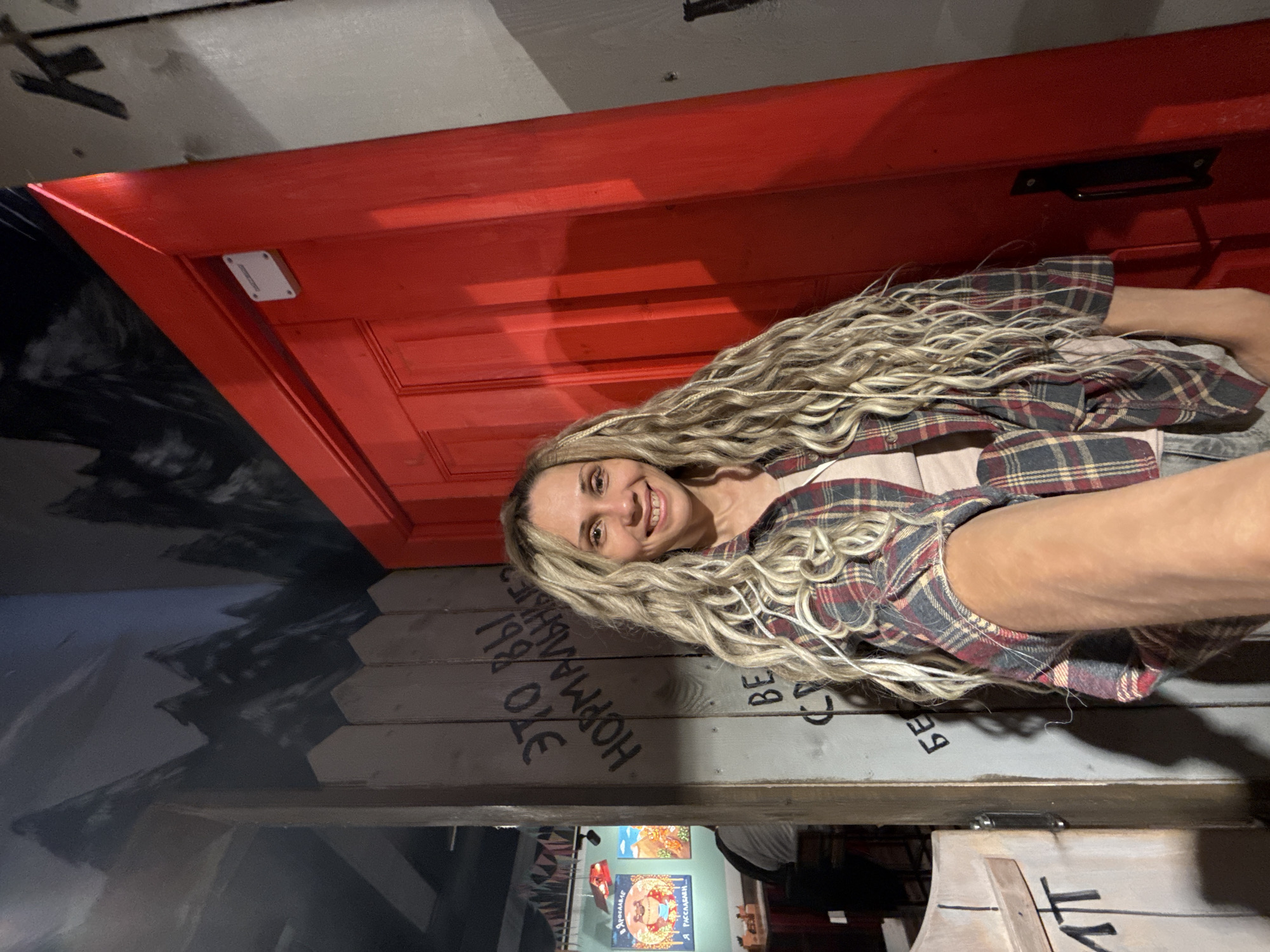 The image, rotated 90 degrees clockwise, captures a smiling woman indoors in Заволжье, Russia. She has long, wavy blonde hair and is wearing a plaid shirt over a lighter-colored top. Her expression is cheerful as she looks directly at the camera, seemingly taking a selfie or being photographed.
Behind the woman is a vibrant red door with paneling, adorned with a small white rectangular device near the top left corner. Below the door, a wall is constructed from horizontal wooden planks, upon which various Cyrillic words are stenciled or painted. Prominently visible are "ЭТО НОРМАЛЬНО?" (Is this normal?) and "Я 1980 ОЗЕ" (I am 1980 Oze), along with fragmented letters like "БЕ" and "CA".
To the left of the woman, a wall is painted with an abstract, dark grey and black design resembling jagged mountains or peaks. Through an opening formed by the wooden planks, a glimpse of a further interior space is visible. This area contains at least two framed posters or pictures on a teal-colored wall, with one clearly showing the text "Я ДУМАЛА" (I thought) and another "Я РАССЛАБЛЮСЬ" (I will relax). The overall setting suggests a themed environment, possibly an escape room, a quirky cafe, or an entertainment venue, given the painted walls and scattered text. The lighting appears artificial, indicating an indoor setting.