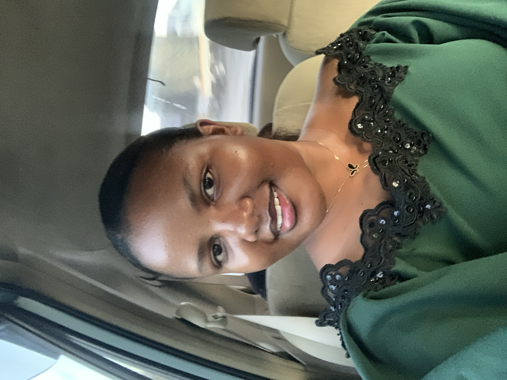 The image is a selfie of a woman sitting in a car. The woman has dark skin and short black hair. She is wearing a green dress with black lace trim and a gold necklace. She is smiling and looking at the camera. The interior of the car is beige. The location is unknown, it is not possible to identify it as Dar es-Salaam, Tanzania.