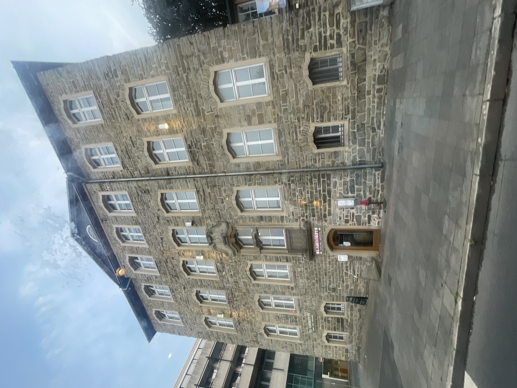 The image shows a multi-story stone building in Iserlohn, Germany, with a restaurant called "EL AMBIENTE" located on the ground floor. The building has a traditional stone facade with multiple arched windows and a dark, sloped roof. The sidewalk in front of the building is paved with gray square tiles. A red fire hydrant is visible next to the restaurant entrance. To the left of the building, a modern gray concrete structure with balconies is partially visible. The sky above is overcast with some clouds.