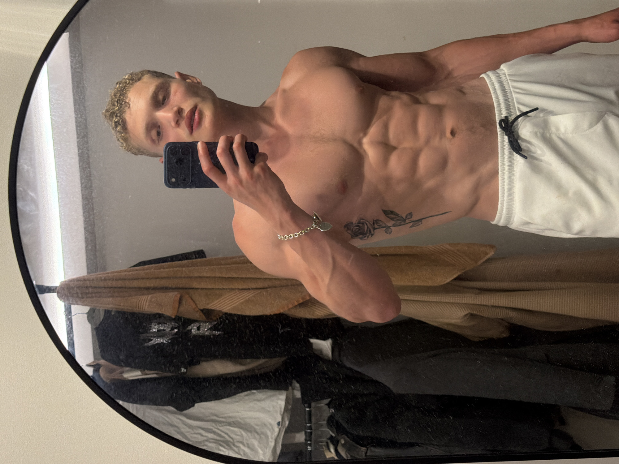 A shirtless, muscular man with short blonde hair is holding a phone in his right hand and taking a selfie in a mirror. He is wearing white shorts with a black drawstring. He has a rose tattoo on his left rib cage and a silver bracelet on his left wrist. The mirror shows a reflection of clothing, including a brown sweater and black items, hanging behind the person taking the picture. The location is likely an indoor room with a white wall. The image does not appear to be set in London, United Kingdom.
