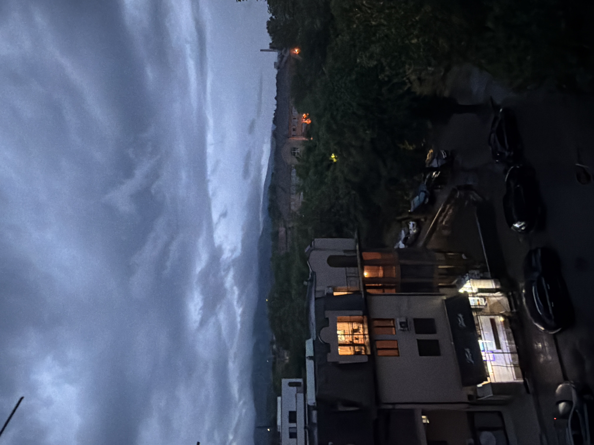 The image captures an evening or nighttime scene from an elevated perspective, looking down onto a street and buildings with a backdrop of hills and a cloudy sky. The overall color palette is dominated by dark blues, greys, and greens, with warm artificial lights providing contrast.
The sky, occupying the upper left and central portions of the frame (when viewed horizontally), is heavily overcast with dark grey and blue clouds, suggesting dusk, night, or inclement weather. Faint outlines of distant mountains or hills are silhouetted against this dim sky.
In the midground, a dense line of dark green trees and foliage spans horizontally across the image, likely indicating a hillside or park area. Within and above these trees, several small, bright orange and yellow lights are visible, suggesting distant buildings or light sources. One prominent, slender, dark structure, possibly a chimney or tower, rises above the tree line on the left. Further right, a larger building with glowing windows is faintly visible on a distant slope.
The foreground and mid-right feature several multi-story buildings. Many windows are illuminated with a warm, golden light, creating a sense of coziness and activity within. One building shows distinct window frames and several air conditioning units mounted on its exterior. Adjacent to these buildings, a street or paved area is visible, with several dark cars parked along its edge. The ground surface appears somewhat reflective, possibly from recent rain or dampness, catching the glow of nearby lights.
The scene conveys a quiet, atmospheric mood, characteristic of an urban or semi-urban setting after sunset. There are no visible people or discernible text within the image.