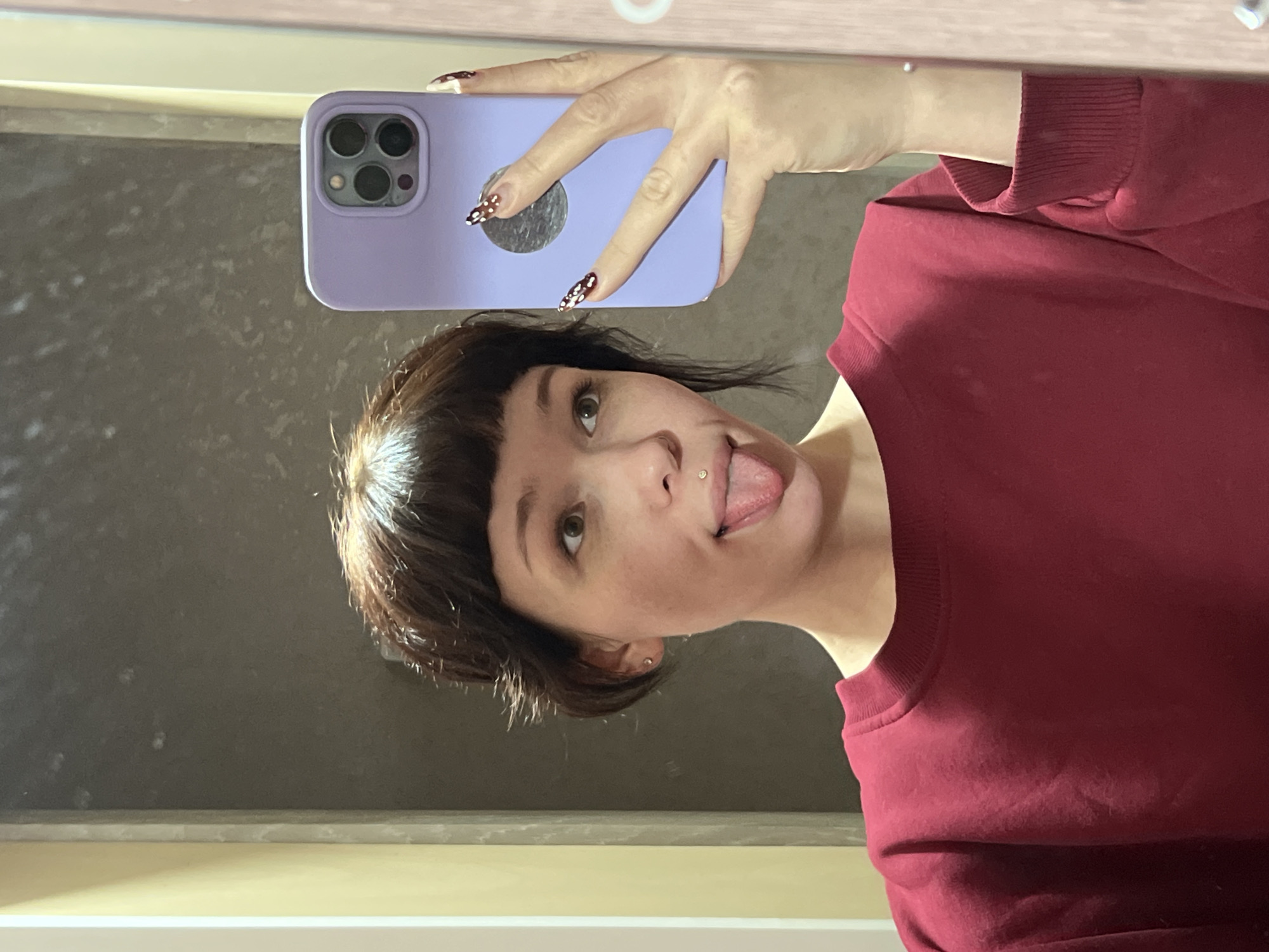 A person with short brown hair and bangs, wearing a maroon sweatshirt, is taking a selfie with a light purple phone case. The person is sticking their tongue out and has a piercing on their upper lip. The nails of the hand holding the phone are decorated with a black and white pattern. The background appears to be a bathroom mirror. The location cannot be determined from the image.