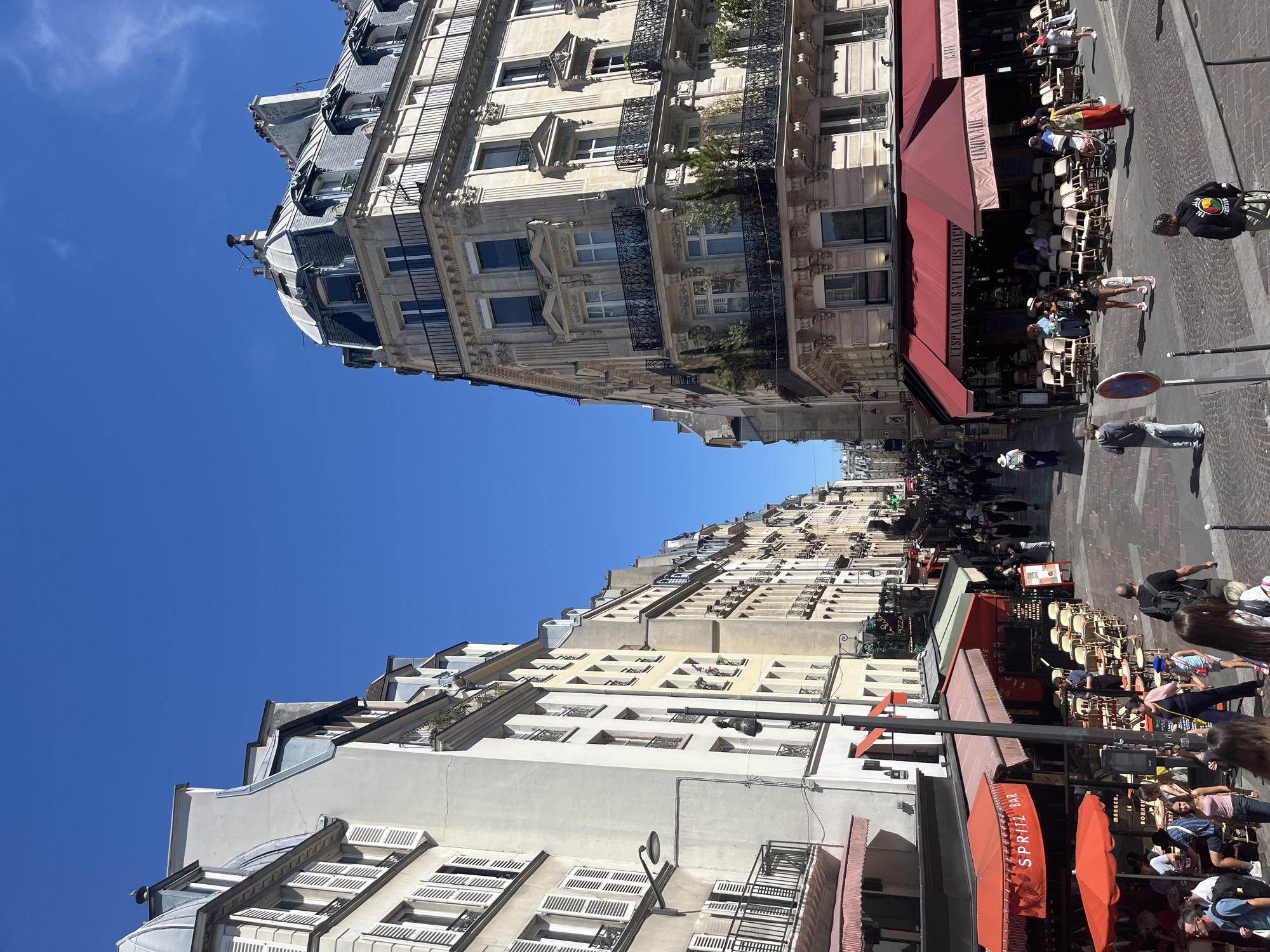 The image depicts a street scene in Paris, France, likely a busy commercial area with outdoor dining. The architecture is classic Parisian, with Haussmann-style buildings featuring ornate facades, mansard roofs, and wrought-iron balconies. The street is paved with cobblestones.
On the left side of the street, several buildings house cafes with red awnings. One awning displays "SPRITZ BAR". Tables and chairs are set up outside, some occupied by patrons. A lamppost extends from a building.
On the right side of the street, a larger building with a prominent domed mansard roof is visible. It also has multiple stories with balconies and windows. Another cafe, named "L'ESPLANADE SAINT EUSTACHE", has a large red awning and tables and chairs set up outside. Signs on this awning advertise "LIMONADE" and "CAFE".
Numerous people are walking along the street and seated at outdoor cafes. Some are dressed casually, while others appear to be tourists. The sky is a clear, bright blue. The scene suggests a sunny day, likely in the spring or summer, and a lively atmosphere. The presence of the cafe signs and the architectural style strongly indicates a location within the historical center of Paris.