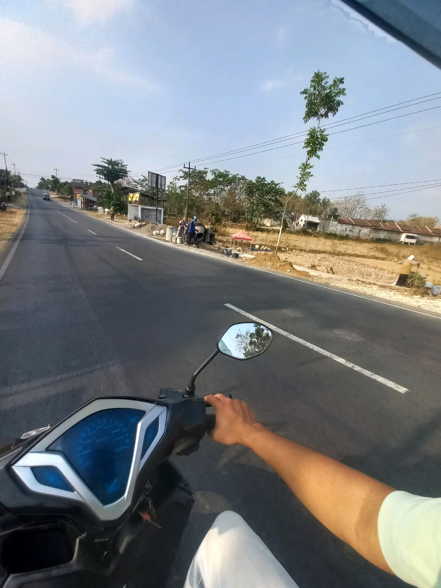 An over-the-shoulder, first-person perspective shot captures a rider's hand on the handlebars of a dark-colored scooter as it travels along a two-lane asphalt road. The road curves to the left and leads uphill into the distance, bordered by dry, barren fields and a few scattered trees and buildings on the right. On the left side of the road, a few more trees and a small building are visible. The sky is clear and pale blue with a few wispy clouds. A single, tall, thin tree with sparse green leaves grows near the middle of the frame, its branches reaching towards power lines overhead. In the background, a white building with a red-tiled roof is visible, along with several other structures. Several people are seen near the side of the road, some appearing to be working with construction materials. One person in a yellow shirt is visible in the middle ground, next to a pile of white buckets. On the far right, two figures in blue shirts and headscarves are interacting. A single vehicle is visible in the distance on the road. The image is taken during the day, likely in the late afternoon, judging by the angle of the sun and the long shadows. The location is specified as Sendangrejo, Indonesia.