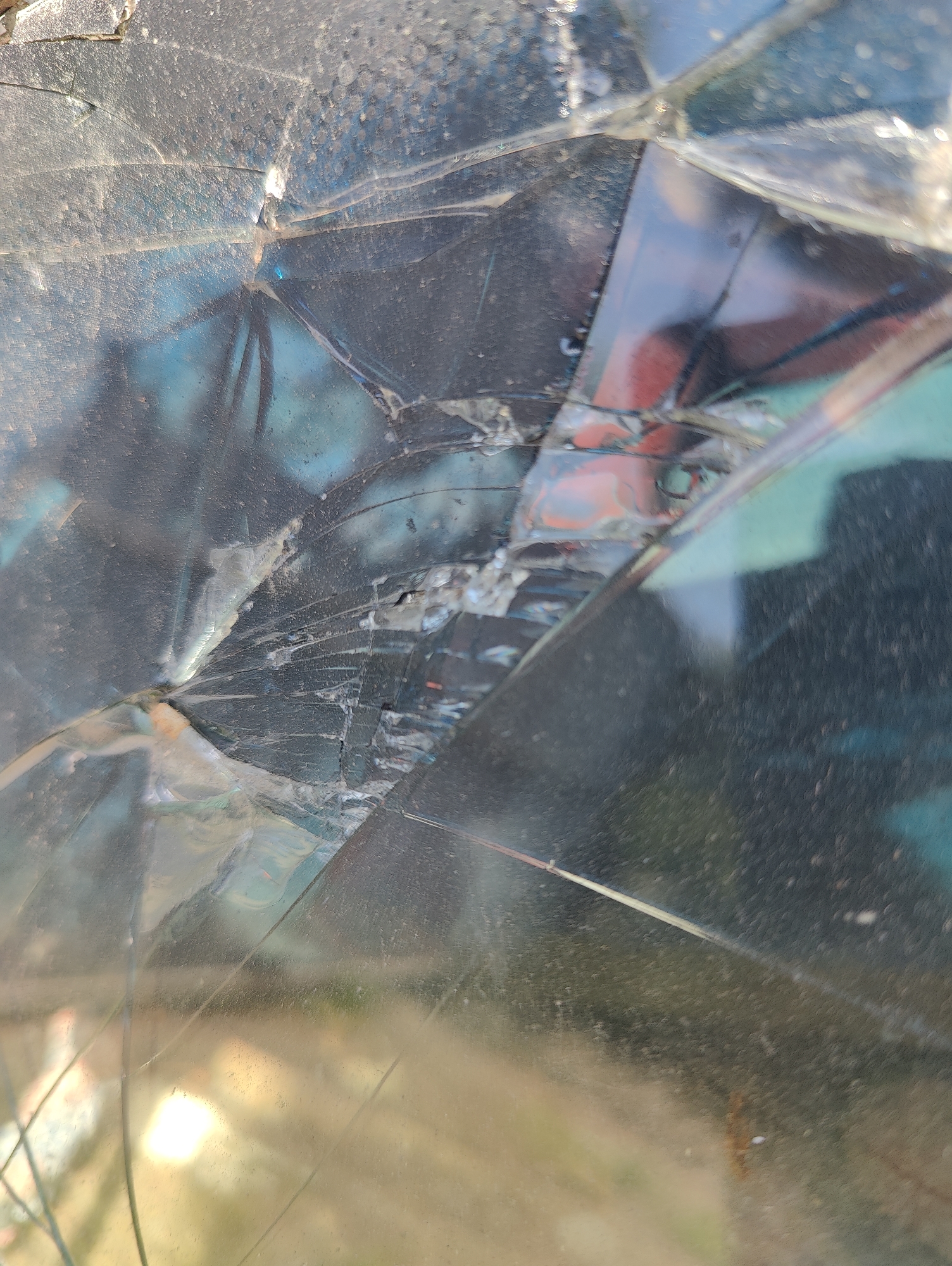 The image is a close-up, abstract view of shattered glass, likely a broken windshield. Cracks spiderweb across the surface, distorting reflections of the environment behind it. Hints of blue and red are visible within the reflections, suggesting the presence of other vehicles or structures. The glass is dusty and smudged, indicating it has been exposed to the elements. The background appears to be an outdoor environment, with muted colors of brown and green visible through the fragmented panes. Without additional context, it is impossible to determine the specific location in Yarmolyntsi, Ukraine.