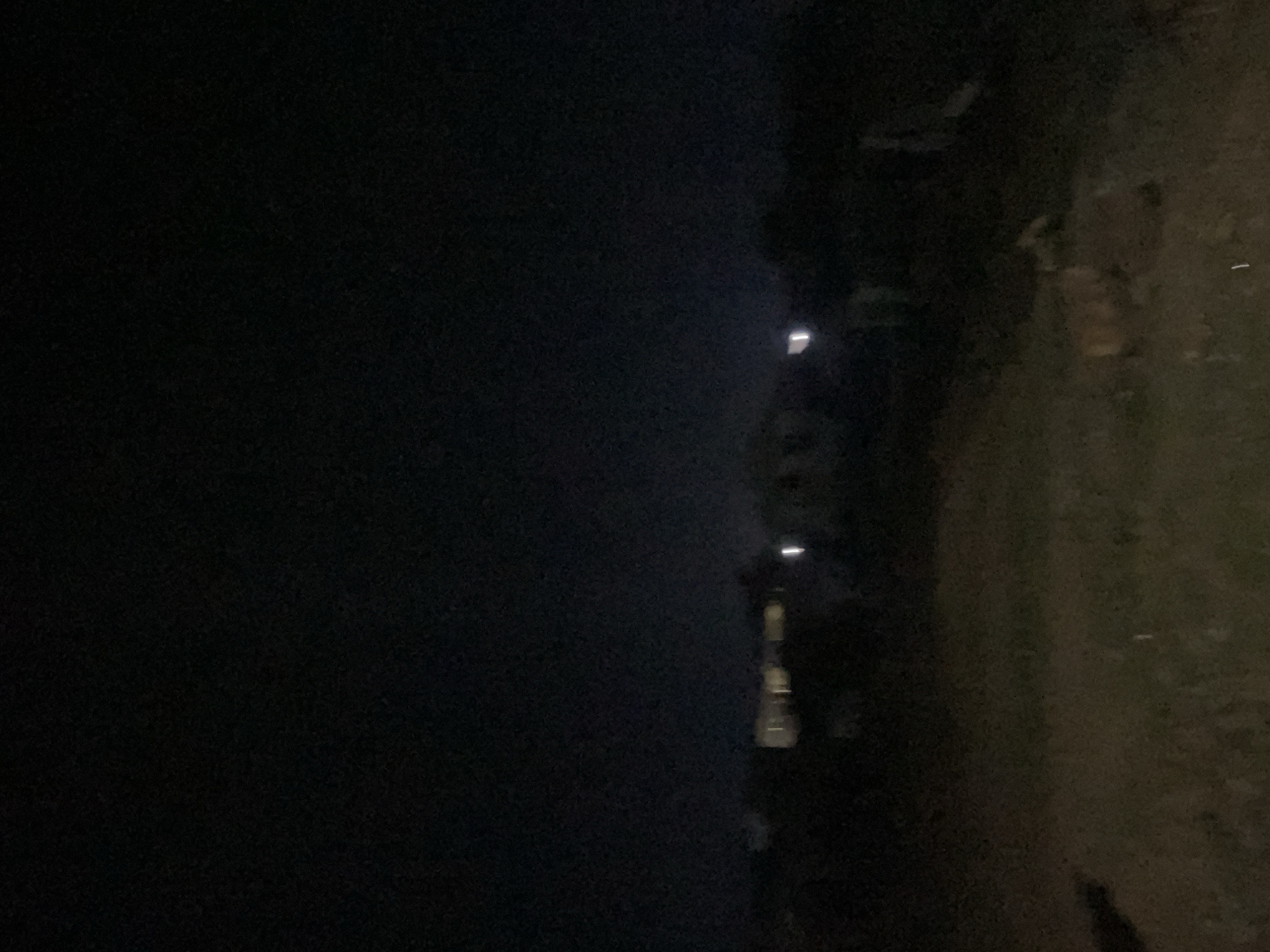 A low-light, wide-angle outdoor photograph captures a dark, rural landscape under a black sky. In the middle ground, several structures with illuminated windows are visible, suggesting buildings. A bright white light, possibly from a streetlamp or a single light fixture, stands out to the right of the structures. The foreground is a dimly lit, uneven terrain, appearing to be unpaved ground with some scattered debris, possibly organic material. The overall impression is one of a quiet, sparsely populated area at night, potentially on the outskirts of Lokoja, Nigeria, given the context.