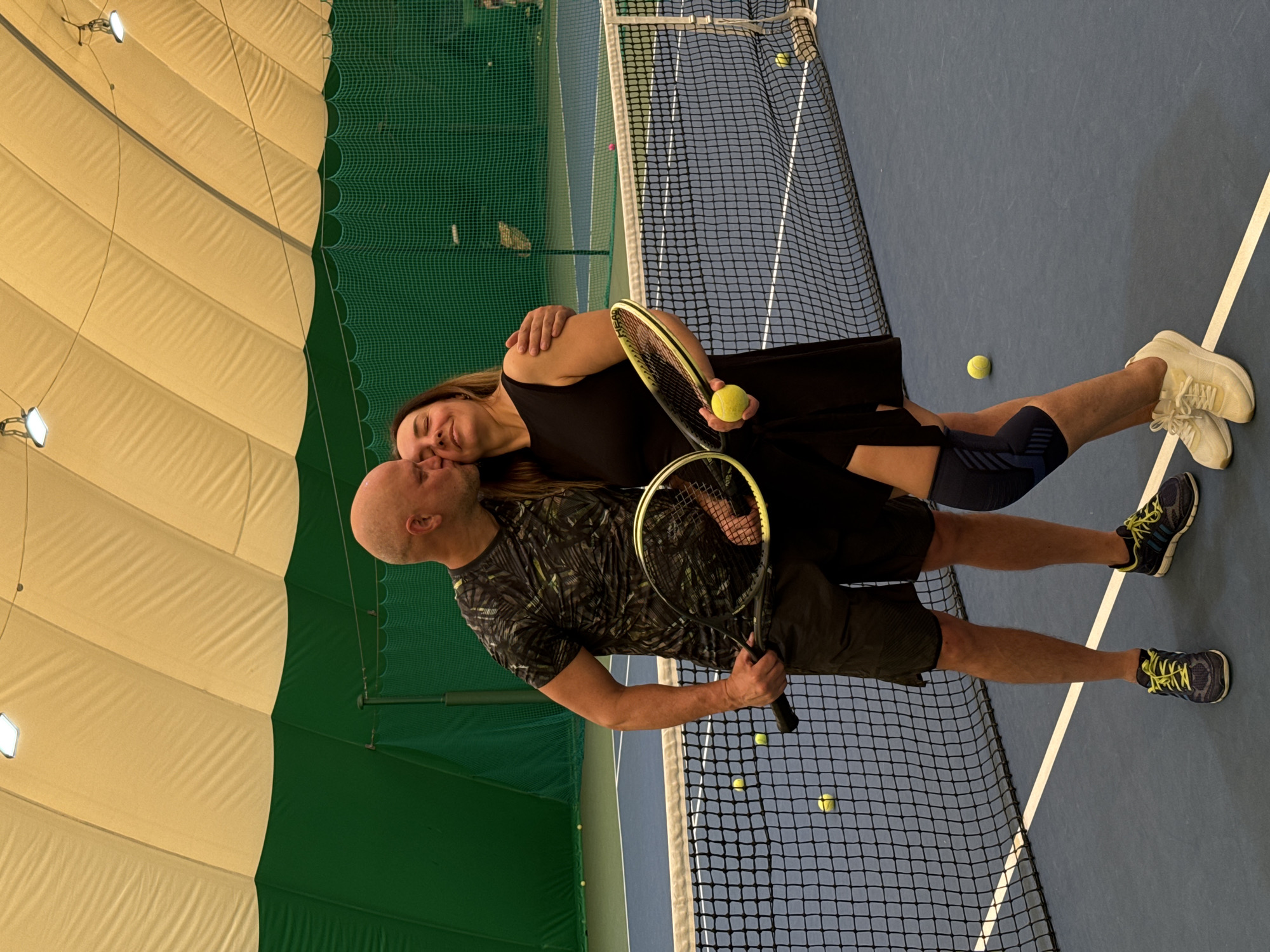 A man and a woman are kissing on an indoor tennis court. The man is bald and wears a patterned shirt and shorts. The woman is wearing a black dress. Each is holding a tennis racket. The court has blue and green surfaces and a net. There are tennis balls scattered around. The roof is constructed of inflated panels. The location is plausibly in the city of Nizhny Novgorod, Russia given the vernacular architecture and the overall cultural context.