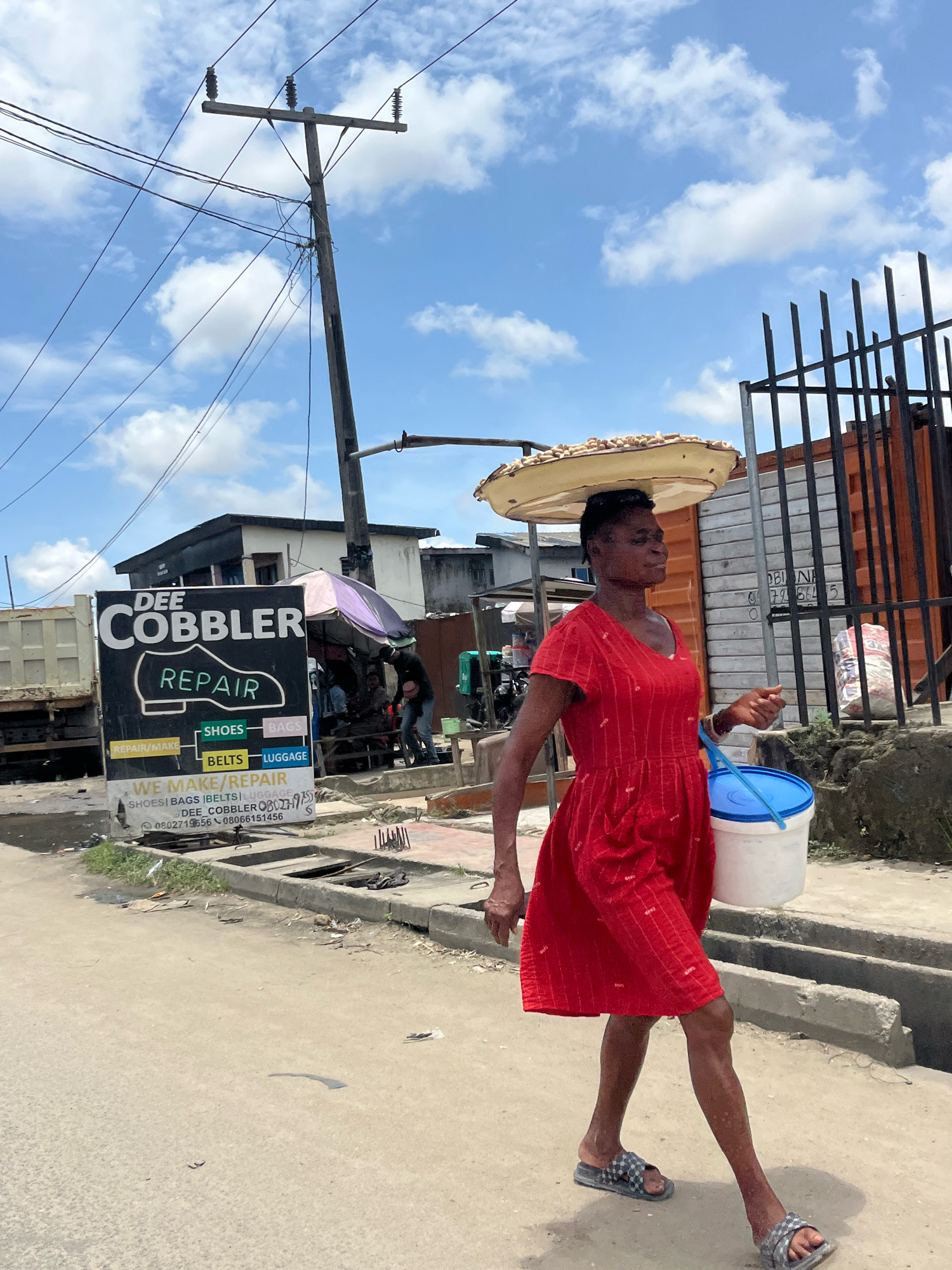 In Ado, Nigeria, a woman walks down a dusty street, her gaze directed ahead. She carries a large, flat tray of what appears to be nuts balanced on her head, her hands free to manage a blue-lidded white bucket in her right hand. She wears a vibrant red V-neck dress with short sleeves, adorned with a subtle repeating pattern. Her feet are clad in grey and black checkered sandals. To her left, a large sign for "DEE COBBLER REPAIR" stands prominently, advertising services for shoes, bags, belts, and luggage, with contact numbers listed. Behind the sign, various structures are visible, including a parked truck, a small shop with a purple and pink umbrella, and what looks like a shipping container. The sky above is a bright blue, dotted with fluffy white clouds, and a wooden utility pole with multiple wires stretches across the scene. The street itself is paved with concrete and shows signs of wear and tear, with drainage channels running alongside.
