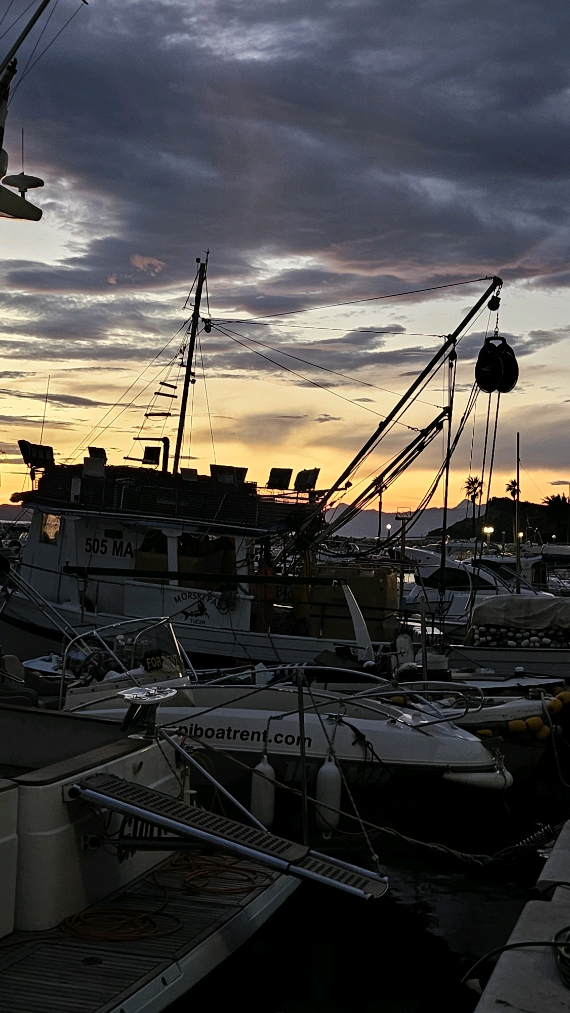 In Općina Tučepi, Croatia, at dusk, a collection of boats are moored in a harbor. A large fishing trawler, marked with "505 MA," is prominently featured in the foreground, its rigging silhouetted against the colorful sky. Smaller vessels are docked alongside it. The sky is dramatic, with dark clouds tinged with orange and yellow from the setting sun. The water is dark and reflects the muted light. The scene captures a moment of calm before nightfall in a coastal town.