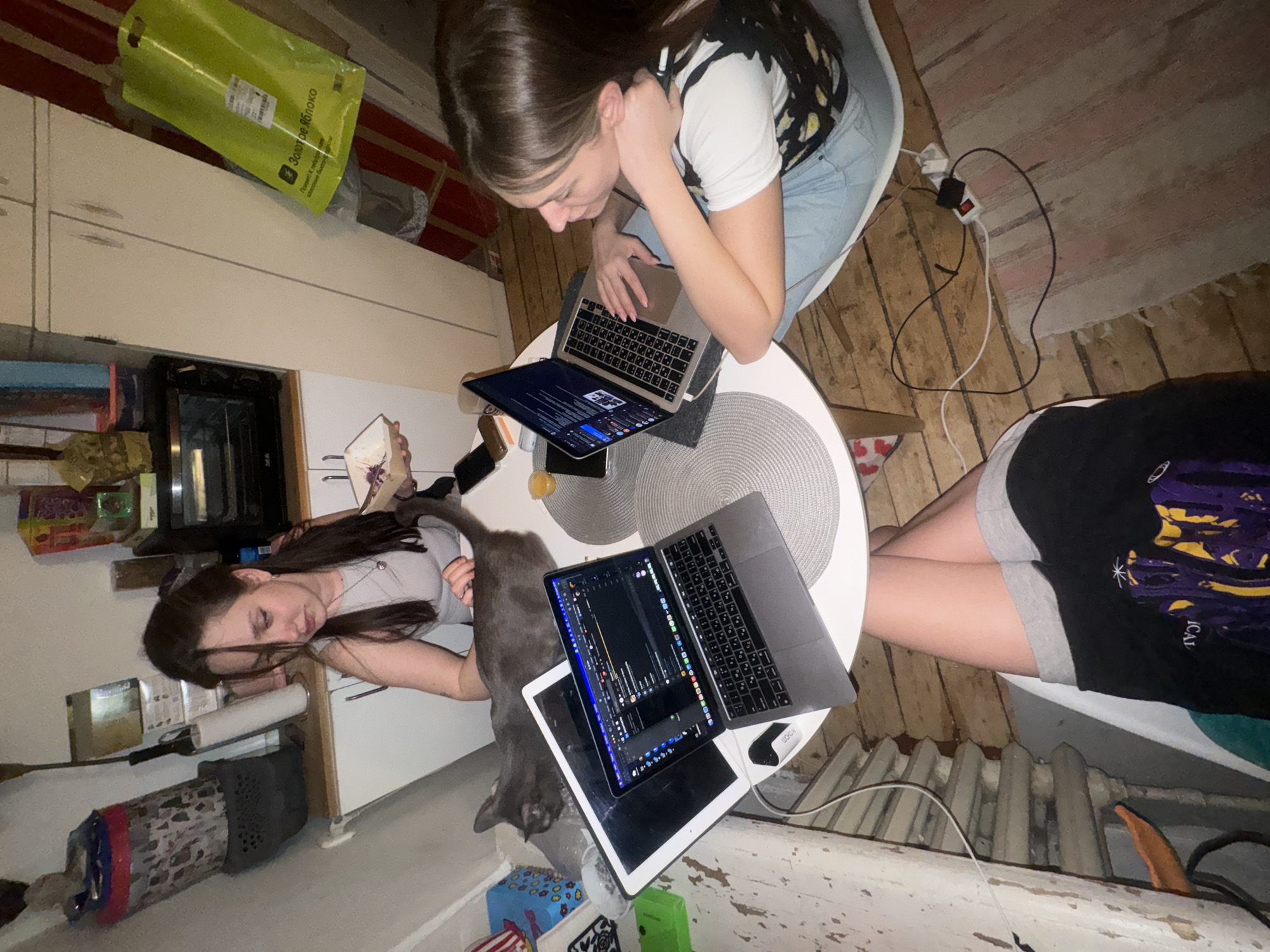 Two young women and a gray cat are gathered around a round white table with a gray woven placemat in what appears to be an apartment in Moscow, Russia. The table holds three laptops and a tablet. One woman is seated at the table, working on a laptop. The other woman is lying down on the floor next to the table. The cat is standing near her. The apartment has a rustic, lived-in feel with wooden floors. An electrical cord with a power strip is near the seated woman. A radiator is visible near one of the women. There is kitchen cabinetry with a yellow bag hanging on the side.
