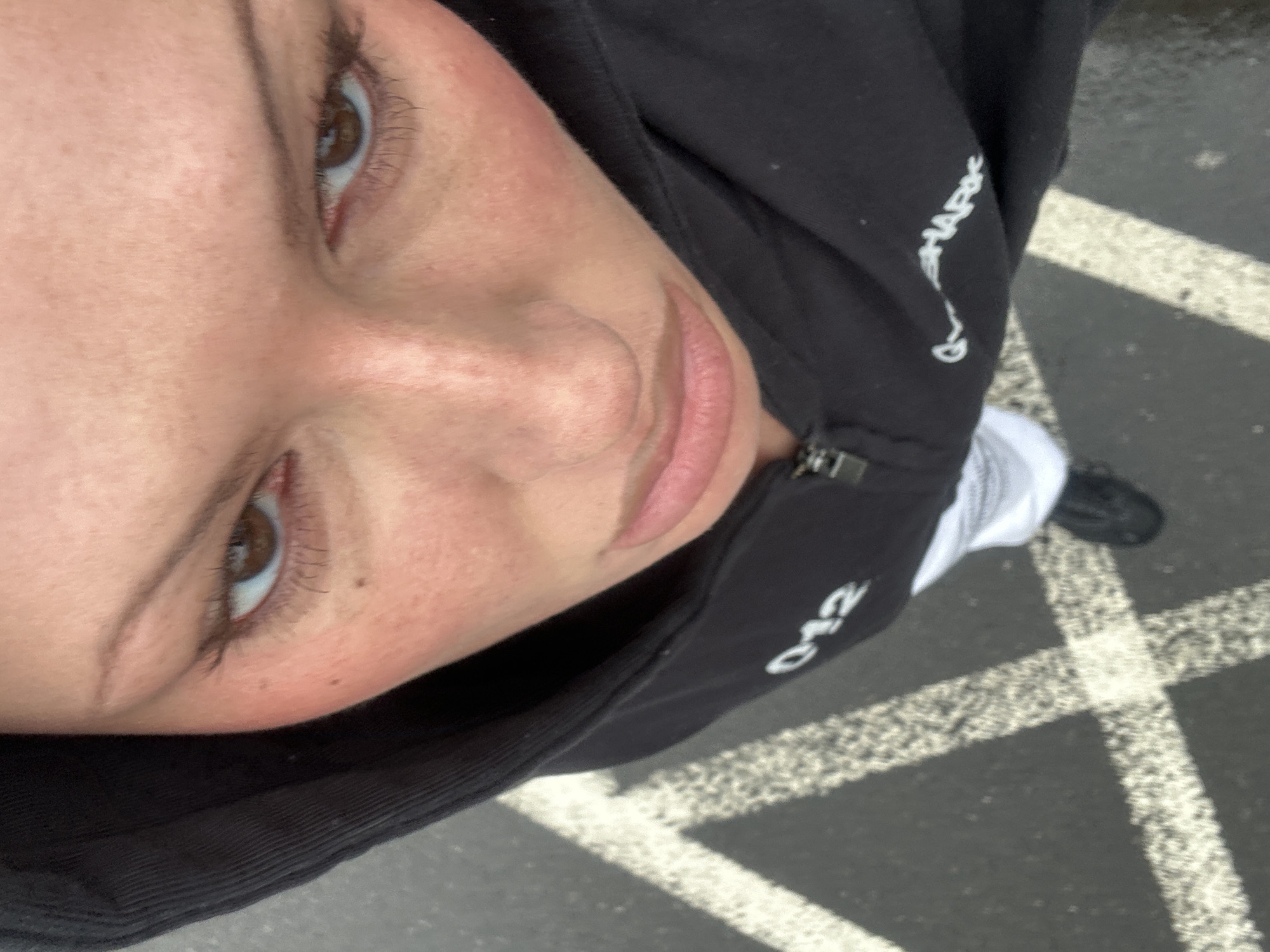 An individual, likely female, is photographed from a high-angle, downward perspective, standing on an asphalt surface. The subject's face features fair skin with some freckles and a reddish complexion on the cheeks. Brown eyes are directed upwards towards the camera, beneath thin, arched eyebrows. The nose is straight, and the lips are full with a slightly downturned appearance. She is wearing a dark, hooded, zippered sweatshirt; the hood is up, partially framing the face. White numbering "012" is visible on the left chest area, and partially visible white text "GYMSHARK" is present on the right side. White trousers or joggers are visible below the sweatshirt, with a dark shoe on the right foot. The asphalt ground displays worn white painted lines, indicative of a parking lot or road marking. The specific location within Tunbridge Wells, United Kingdom, cannot be determined from the visual evidence present in the image.