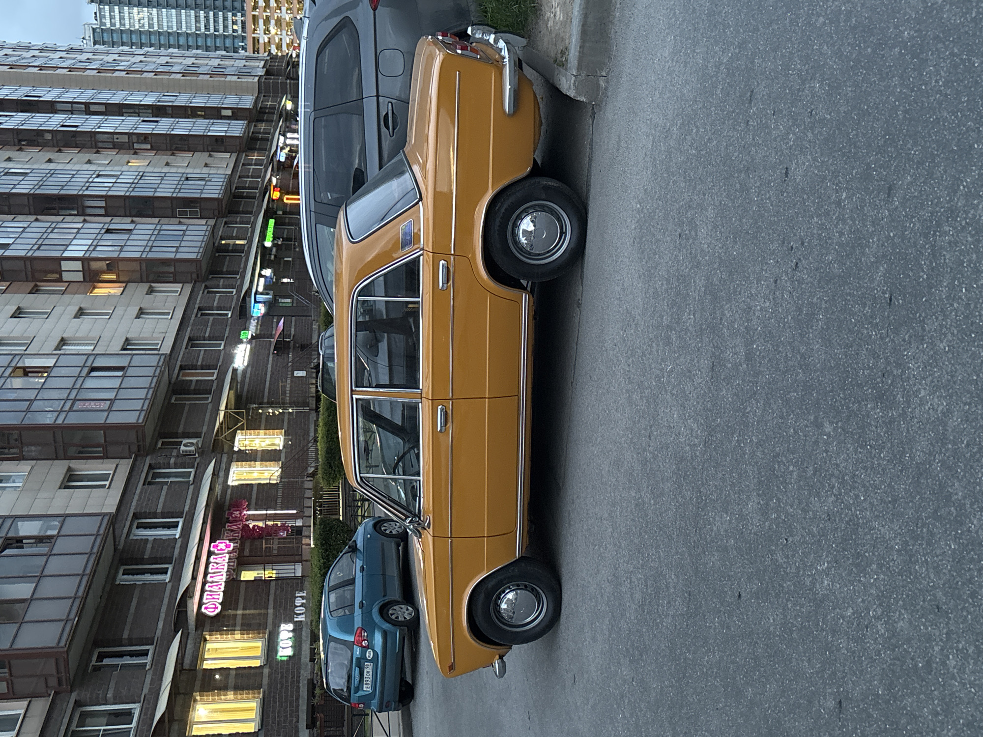 The image depicts a street scene in Saint Petersburg, Russia. A yellow Lada sedan is parked on the right side of the frame, facing away. Adjacent to the Lada is a gray SUV. Behind the Lada, a blue hatchback is visible. A multi-story building is on the left side of the frame. The building features numerous windows and some balconies. There are business signs in Russian on the building. The road in the foreground is asphalt. The scene appears to be during the daytime, possibly late afternoon or early evening.