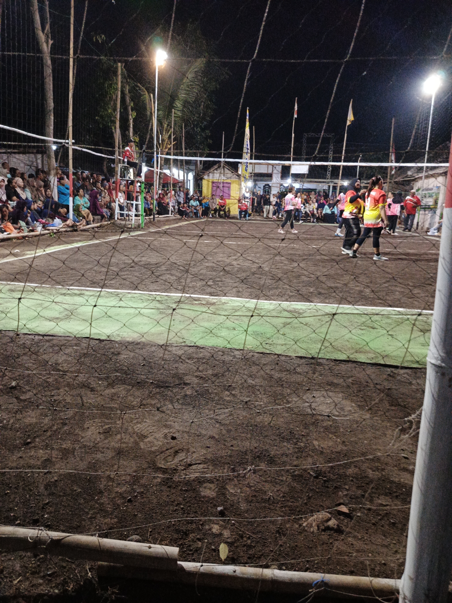 The image depicts a nighttime volleyball game on a dirt court in Rowokangkung, Indonesia. A net divides the court, with players in brightly colored uniforms actively engaged in the match. Spectators are gathered on the side, seated on benches and the ground, watching the game. The scene is illuminated by overhead lights, casting shadows and creating a lively atmosphere. Trees and some structures are visible in the background, suggesting an outdoor setting within the town. The court itself appears to be a well-used, informal space, with the ground showing signs of wear and tear.