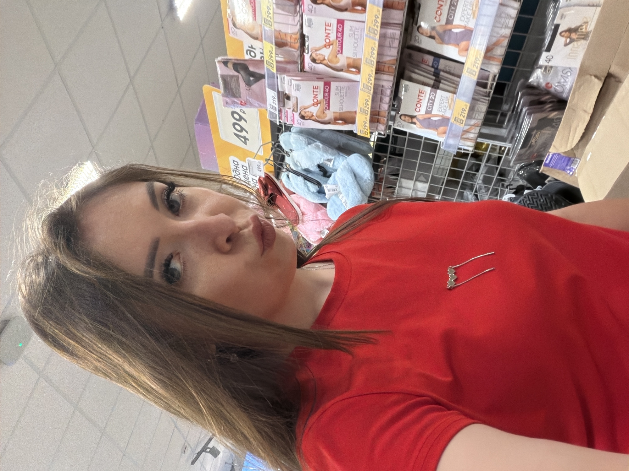 A young woman with long brown hair and full makeup is posing for a selfie in a supermarket aisle in Nurkeevo, Russia. She is wearing a bright red t-shirt with a silver heart-shaped pin on it. The background shows shelves stocked with various products, including what appear to be hosiery and slippers. A price tag with "499" is visible on a yellow sign. The lighting suggests an indoor retail environment.