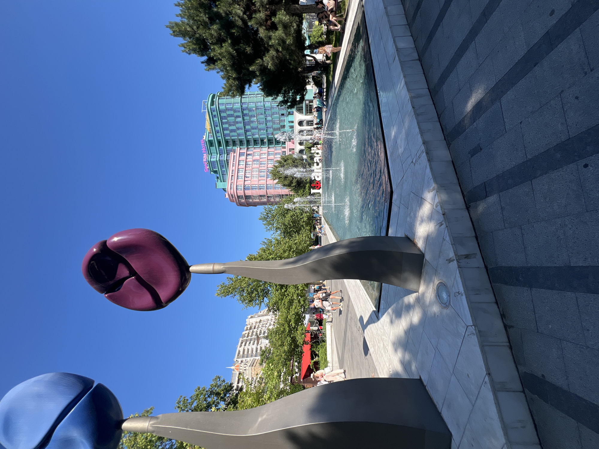 This image depicts the "I love Arcadia" fountain and sculpture in the city of Odesa, Ukraine. In the foreground, two large, stylized tulip sculptures are visible; one is blue and the other is a deep purple. They are placed on metallic stands. A water fountain is active in the center of the image, with several jets of water spraying upwards. The fountain is surrounded by a shallow pool with a light-colored tiled edge. Behind the fountain, the words "I love Arcadia" are spelled out in white block letters, with a red heart emoji integrated into the design.
In the background, several buildings are visible. The most prominent are two brightly colored, modern high-rise hotels, one painted pink and the other teal. The teal building has "SRATIUM HOTEL" written in neon pink letters near the top. Trees line the area, providing shade. People are scattered throughout the scene, some walking along the paved pathway, others seated on benches. The sky is clear and blue, indicating a sunny day. The ground is paved with tiles, with some areas featuring a darker stone.