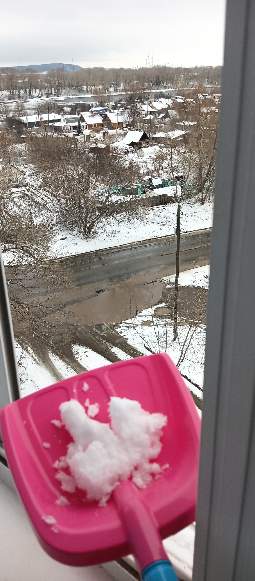 From a window view, a pink plastic shovel filled with snow is in the foreground. The middle ground features a snow-covered street with bare trees and a few puddles of water. In the background, there's a suburban area with houses and snow-dusted roofs, transitioning to a distant landscape with trees and utility poles under an overcast sky.