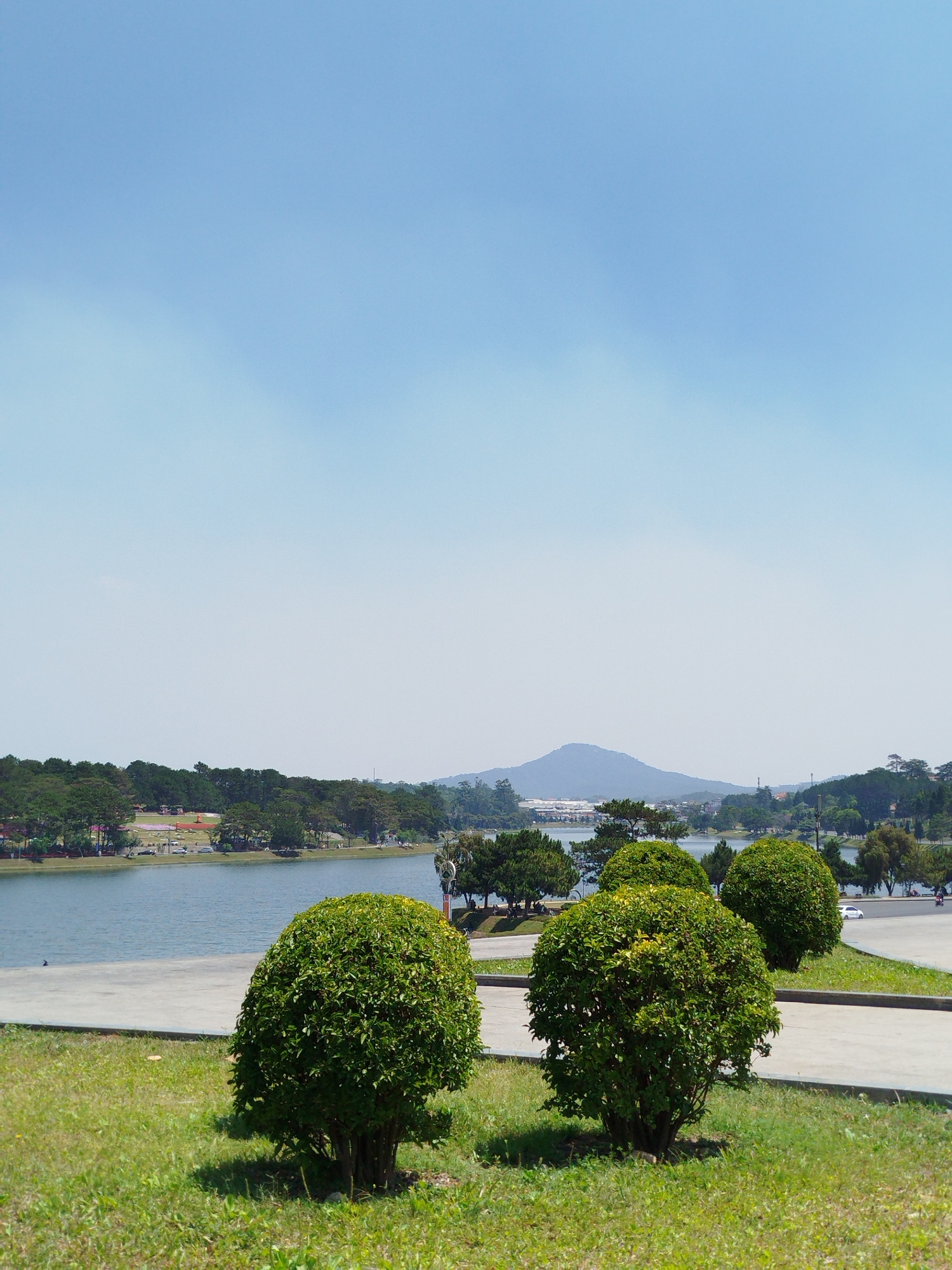 The image captures a sunny daytime view of Xuan Huong Lake in Da Lat, Vietnam. The sky is clear and blue. The lake stretches across the midground, framed by trees and vegetation along its banks. In the foreground, there is a grassy area with three round, trimmed bushes. A paved walkway runs parallel to the lake. In the distance, a mountain can be seen.