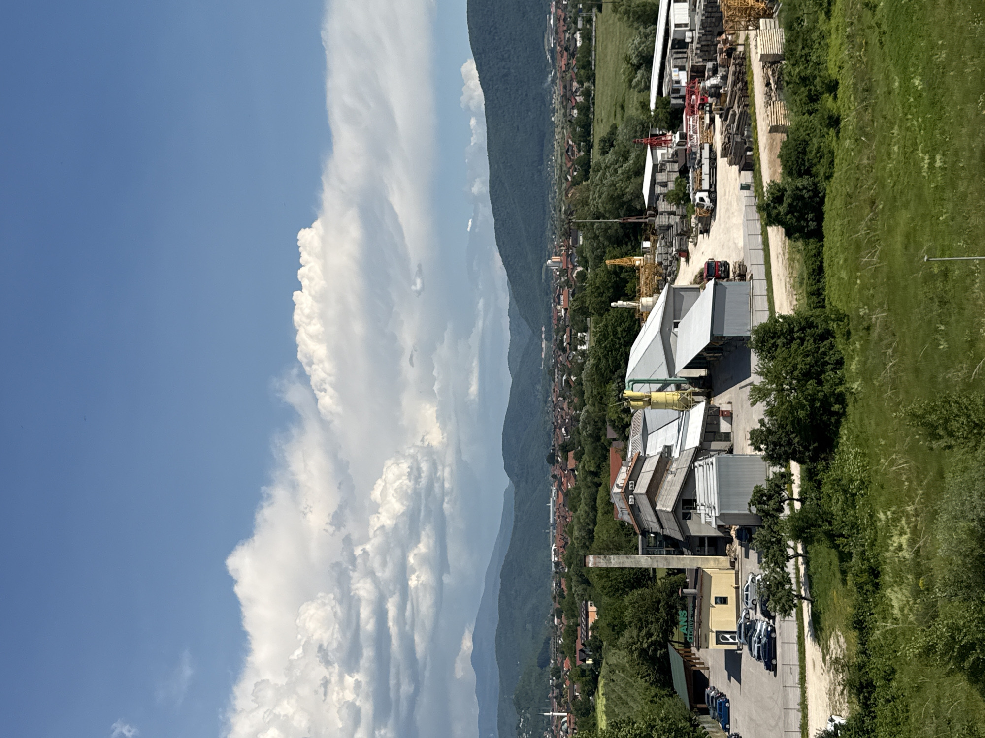 The media file depicts a scenic view of a semi-industrial area nestled beside a lush green hillside, with a majestic mountain range forming the backdrop. The scene is captured from an elevated perspective, likely from a drone or a high vantage point. The sky is a vibrant blue, dotted with fluffy white cumulus clouds, suggesting a bright and fair-weather day.
In the foreground, to the right, a verdant grassy slope dominates, with trees and bushes interspersed. As the view moves left, an industrial complex comes into focus. This complex features several buildings with metal roofs, along with stacks of construction materials such as large concrete slabs and wooden planks. Cranes, both yellow and red, are visible, indicating ongoing construction or operational activities within the site. A dirt road winds through the industrial area, with several vehicles, including cars and what appear to be trucks, parked or moving within the complex.
Beyond the industrial zone, a densely populated residential area is visible, characterized by numerous houses with red-tiled roofs, interspersed with trees. This town stretches along the foothills of the imposing mountains. The mountains themselves are covered in dense green vegetation, with layers of them receding into the distance, creating a sense of depth and scale.
The lighting suggests it is daytime, likely mid-morning or afternoon, given the bright sunlight and clear skies. The overall impression is one of a landscape where industry and nature coexist, with a town situated between them, all under a vast, open sky. The location context is Ilidža, Bosnia and Herzegovina. No people are clearly discernible in the image. One building has visible text that reads "ANS DRIVE".