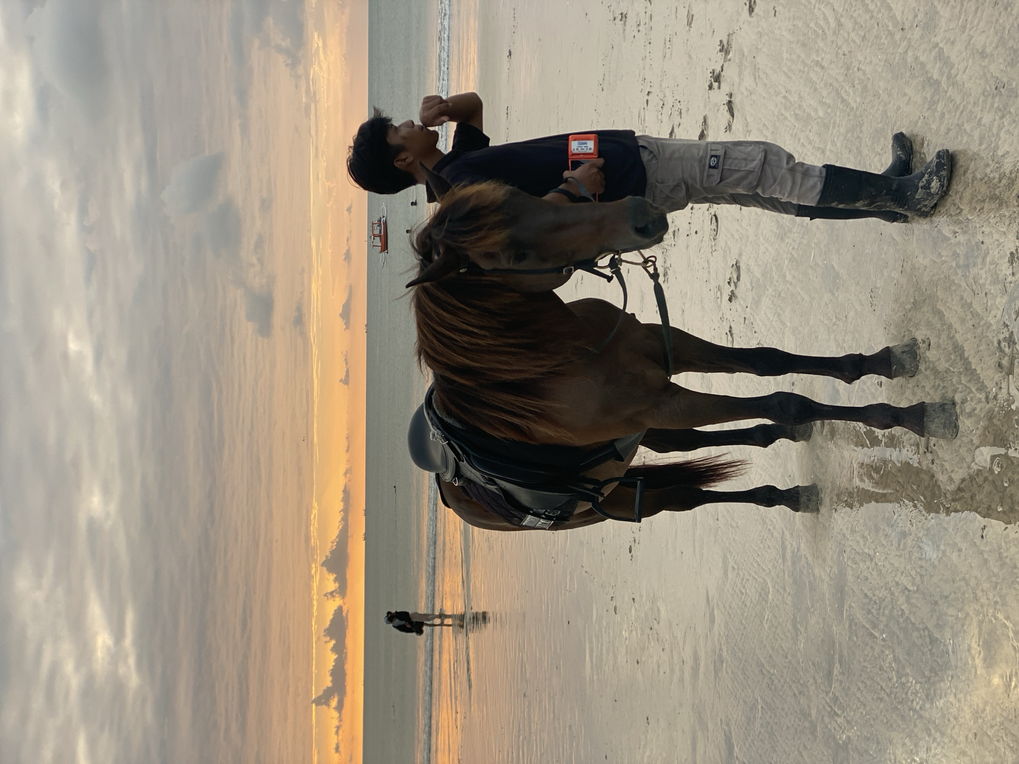 A dark brown horse stands on a wet sandy beach, its head turned slightly to the right. A young man, wearing a dark t-shirt, grey cargo pants, and black boots, stands beside the horse, holding its reins and holding a small orange device in his right hand. The man's left hand is near his mouth. In the background, a person in light-colored clothing stands in the shallow water, facing away from the camera. The ocean stretches to the horizon, with several small boats visible in the distance. The sky is filled with dramatic clouds, illuminated by the warm hues of an approaching sunset or sunrise, casting a golden glow over the scene. The wet sand reflects the sky and the figures, creating a serene and atmospheric beach environment. The location is the beach in Kuta Selatan, Indonesia.