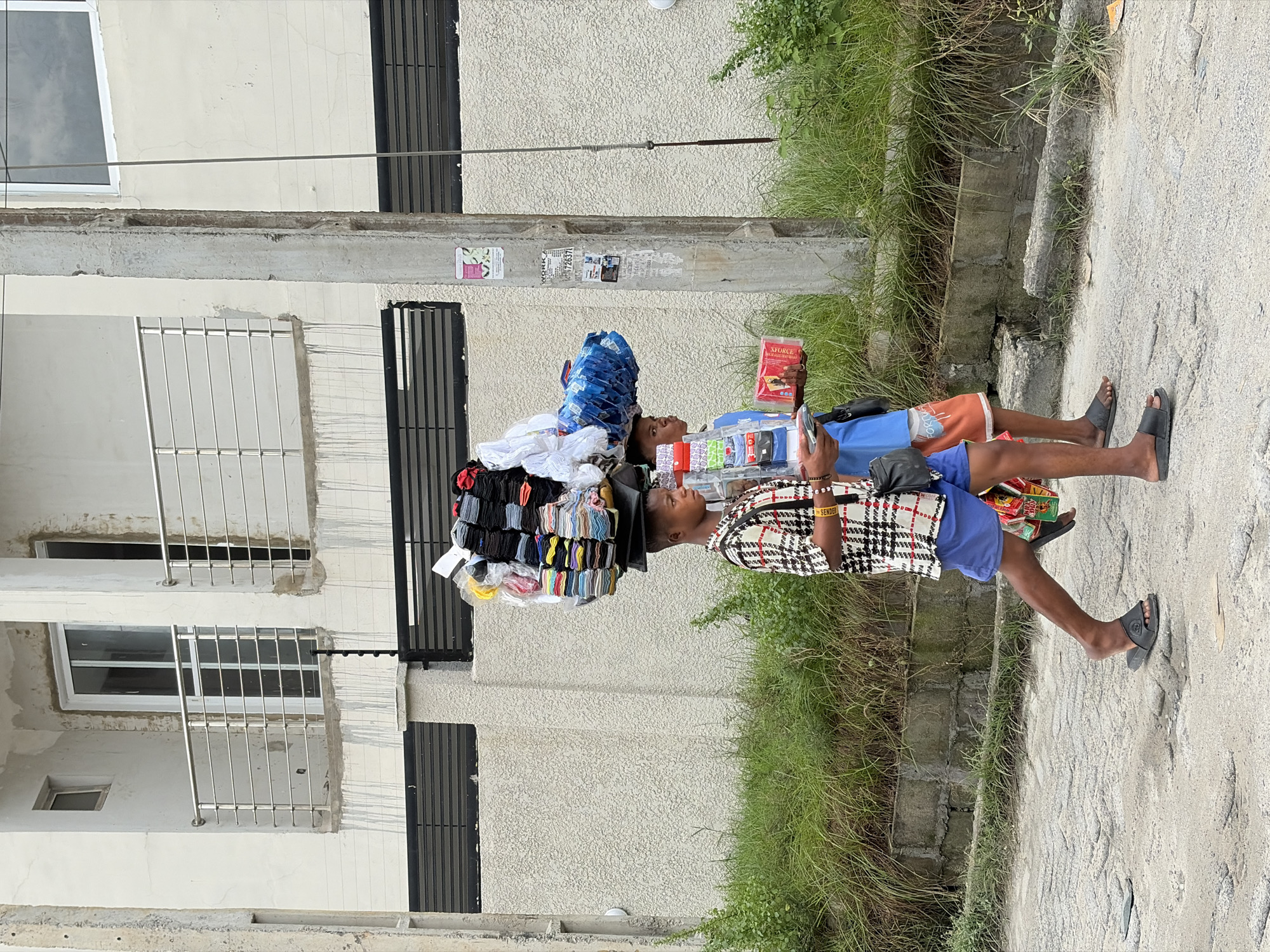 Two young boys are walking on a paved street in Igboefon, Nigeria. The boy on the left is carrying a large stack of items on his head, including what appear to be folded clothes and plastic bags. He is wearing a patterned short-sleeved shirt and blue shorts, and black flip-flops. The boy on the right is carrying a red box in his right hand and a stack of items on his left arm. He is wearing an orange and white patterned shirt and orange shorts, and black flip-flops. The street is lined with overgrown green grass and vegetation. In the background, there is a light-colored building with balconies and railings, and a concrete pillar with posters attached. The sky is overcast.