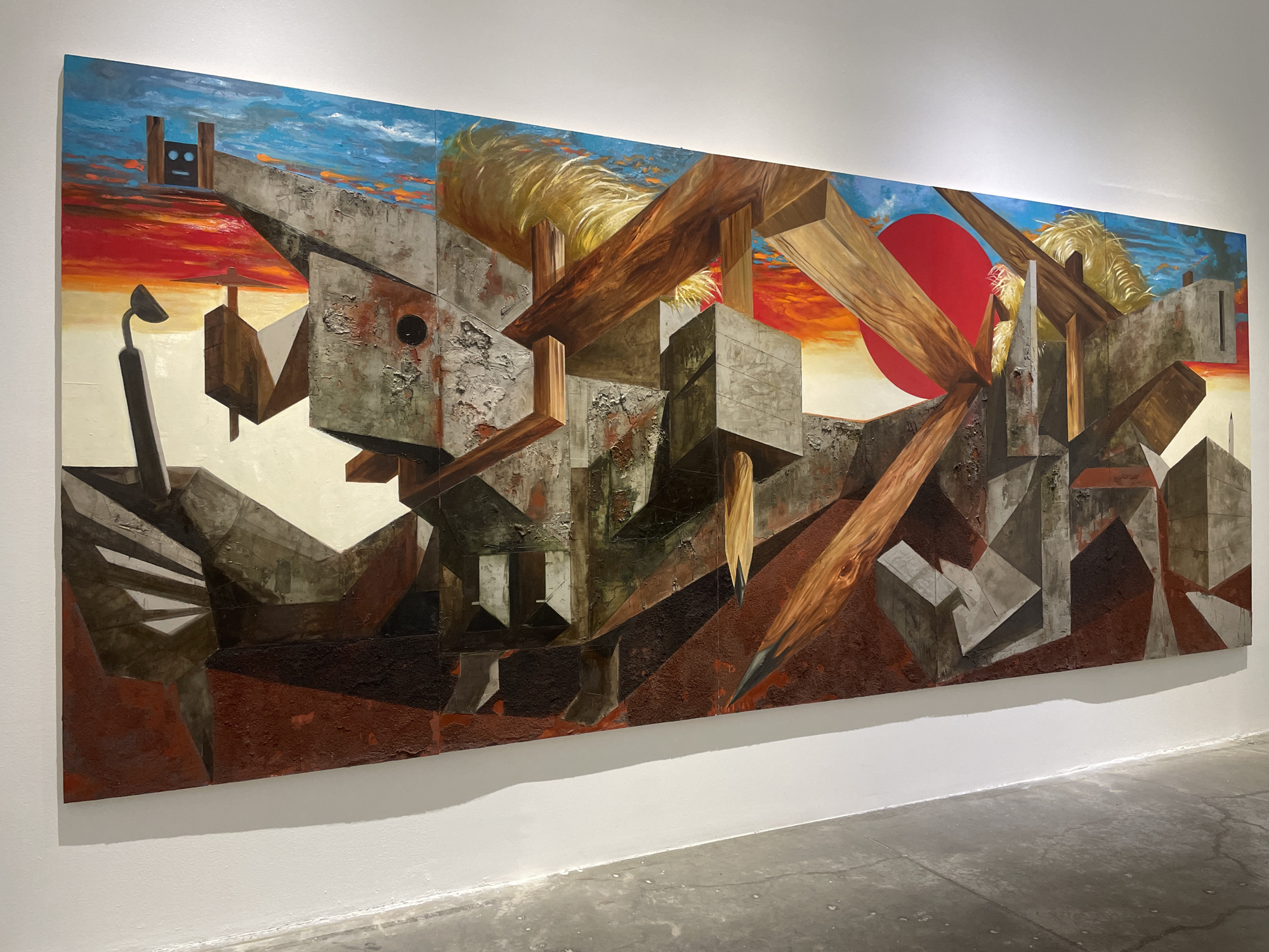 The image shows a large, horizontally-oriented abstract painting hanging on a white wall inside a gallery or museum space. The concrete floor is visible at the bottom, extending from the wall. The painting depicts a complex scene with geometric shapes, including cubes and angular structures, in varying shades of gray and brown, suggestive of concrete and wood. The sky is painted with blue and red hues suggesting a sunset. A red circle is present in the right center. There are hints of yellow straw-like textures. The painting is situated in Mexico City, Mexico.