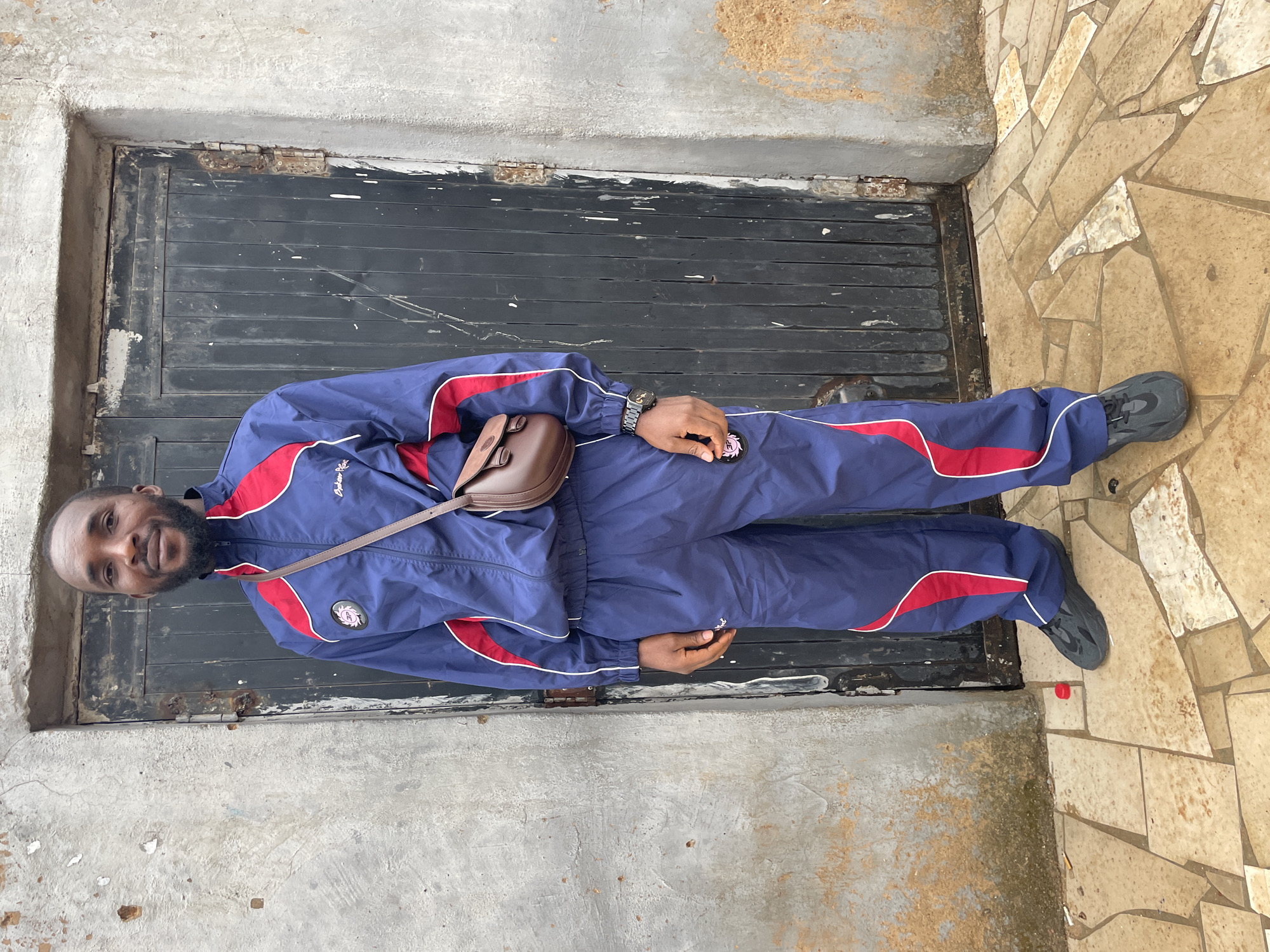 A man stands facing the camera, wearing a blue tracksuit with red stripes, gray sneakers, and a brown shoulder bag. He has dark skin, a beard, and short hair. He is positioned in front of a weathered black door set in a gray concrete wall. To the right of the door is a tiled surface made up of irregularly shaped light brown stones. A red bottle cap sits near his right shoe. The location is possibly a building exterior in Jos, Nigeria, given the subject's likely ethnicity and attire, although there is no specific visual confirmation in the image.
